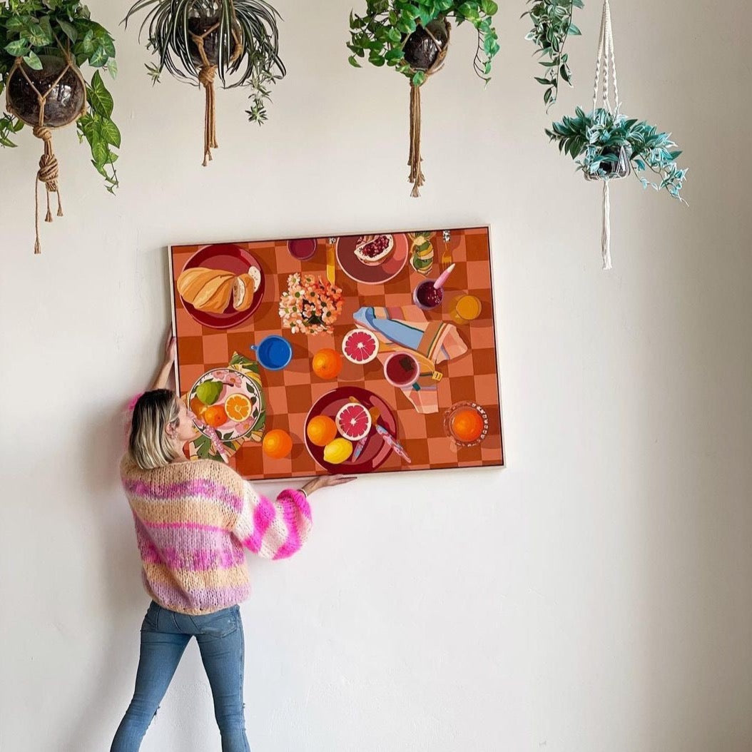 lifestyle photo of Pip from Jumbled in Orange NSW holding a framed canvas print of a breakfast setup with oranges, lemons, tea towels, cutlery by kip and co, and flowers and cups on a chequered tablecloth background