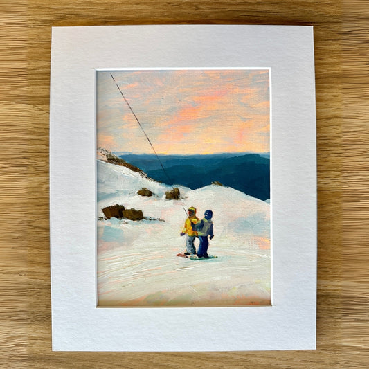 Framed artwork of two people on skis at sunset with a white mat on a wooden surface