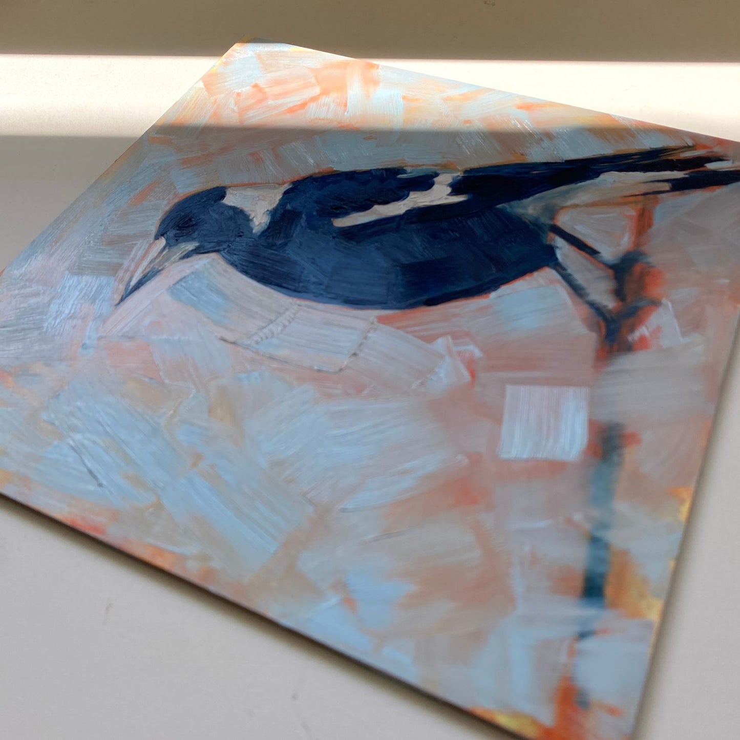 original oil painting of a navy blue and white magpie on a textured sky blue background where you can see orange coming through. The painting is on a cream coloured desk