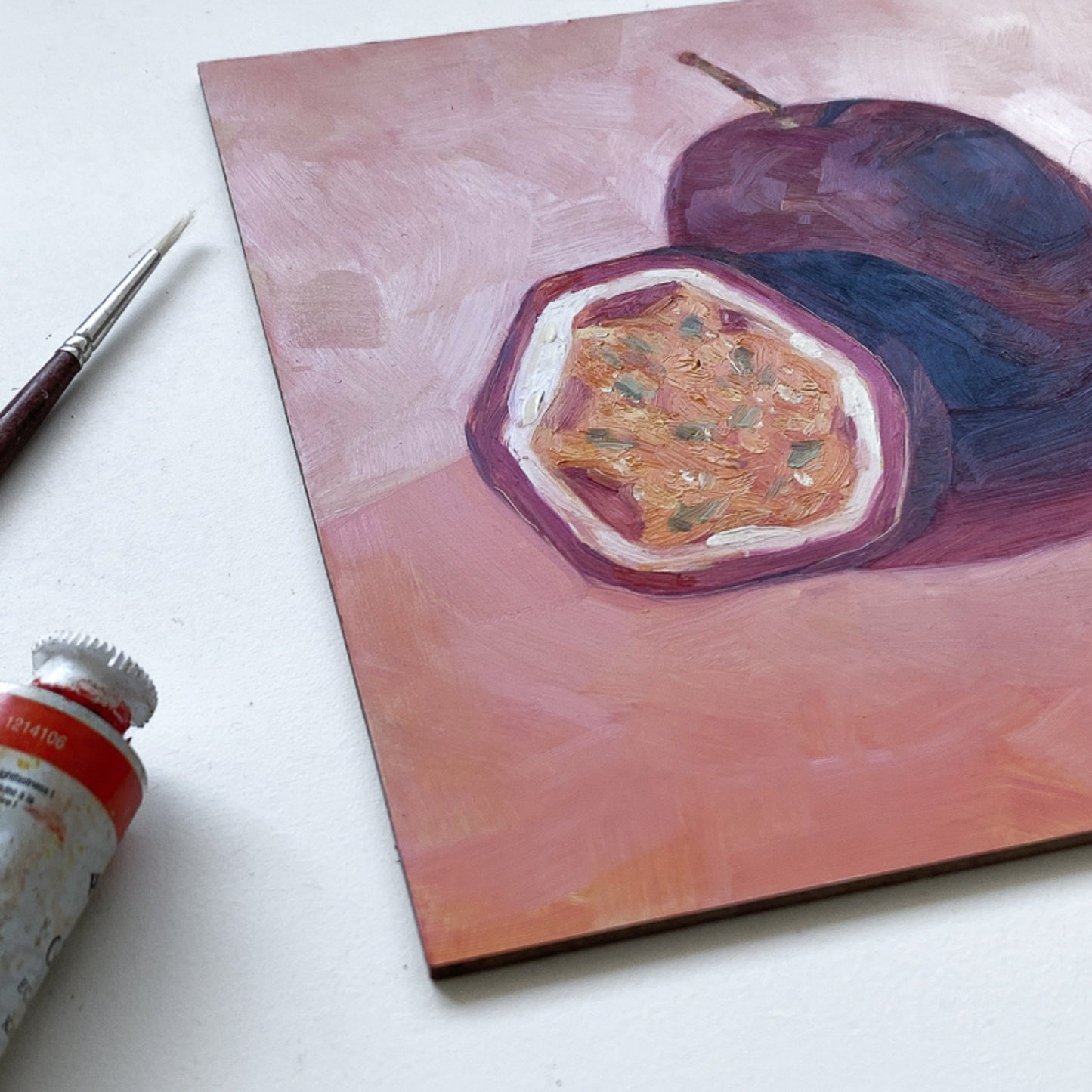 styling photo of an original painting of a burgundy, magenta whole passionfruit and half a passionfruit with seeds. They are sitting on a soft pink surface and there are strong shadows and a diagonal background. The painting is on a white surface and there is a paintbrush and oil paint tube next to it.