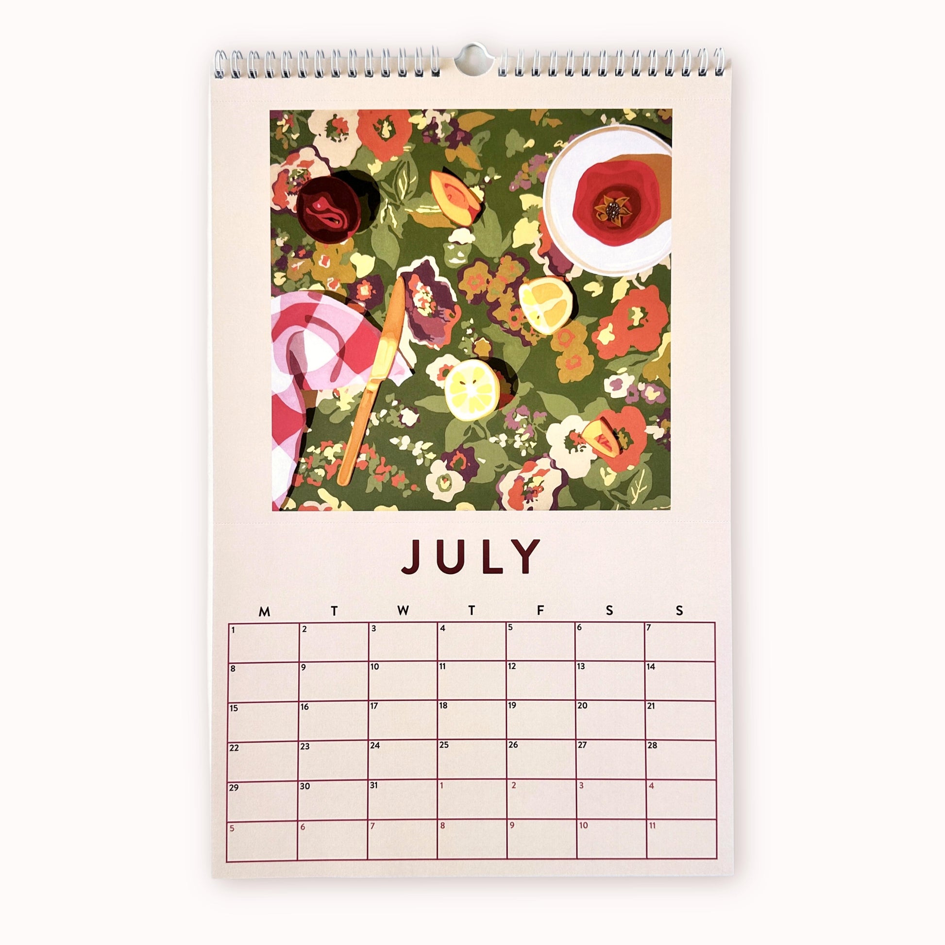 2024 art wall calendar in A3 size for the month of July, featuring a still life oil painting of fruits such as pomegranates with seeds, plum, lemon slices and peach slices, and a chequered tea towel with a knife on a floral green, purple, coral orange mustard patterned background