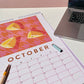 lifestyle photo of a 2024 art wall calendar back cover in A3 size featuring an original oil painting artwork on a desk with an open pen and a laptop apple Mac computer