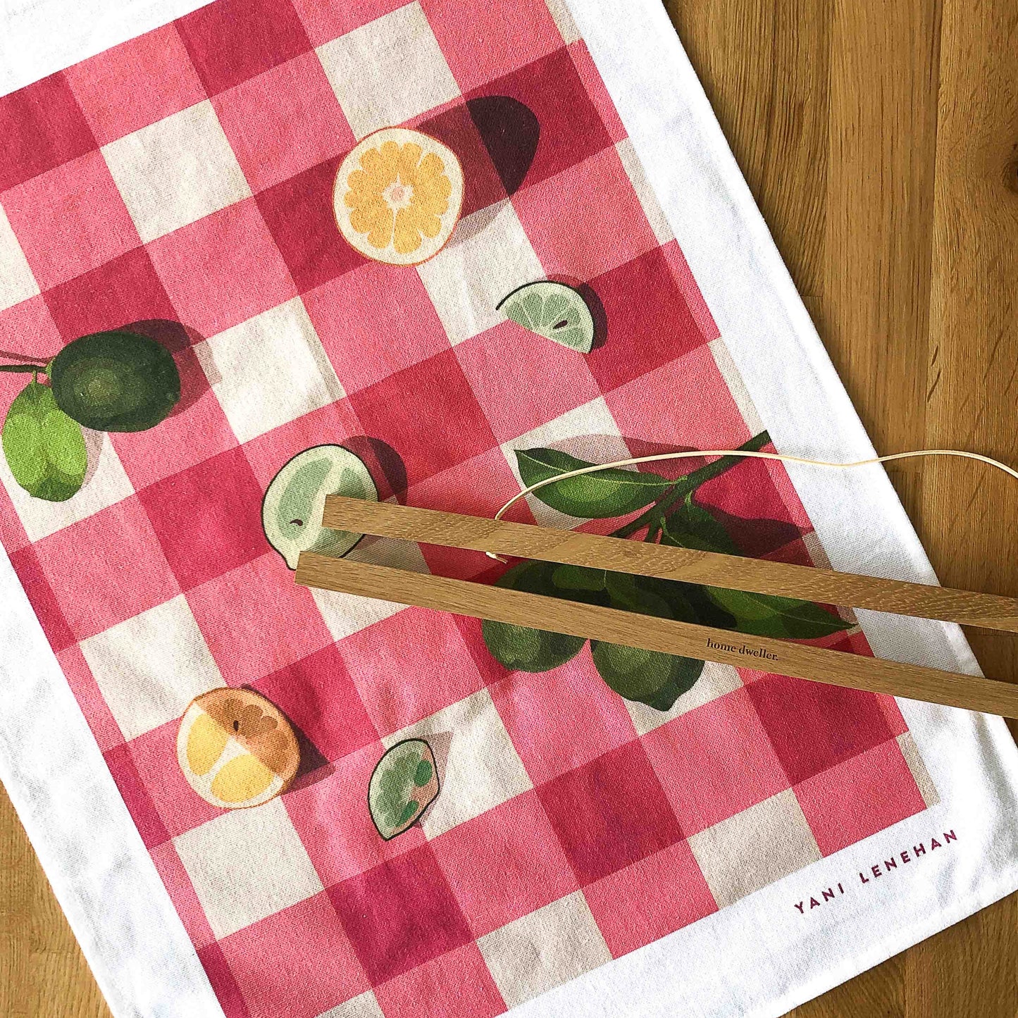 photo of an art tea towel with lemons and limes on a pink-red gingham background on a timber table with wooden magnetic hangers on top