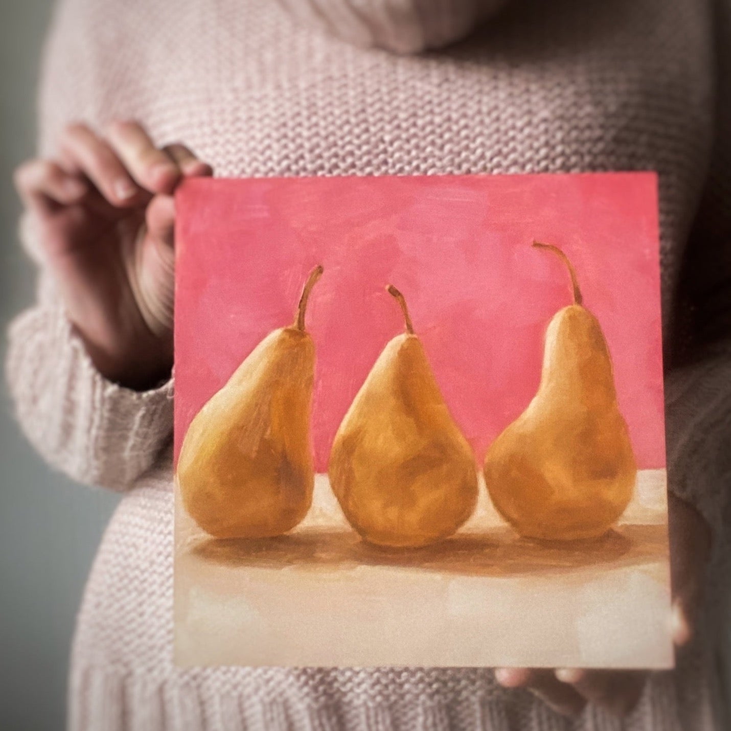 image of a person holding an original oil paintings of three beurre Bosc pears on a warm pink and creamy background