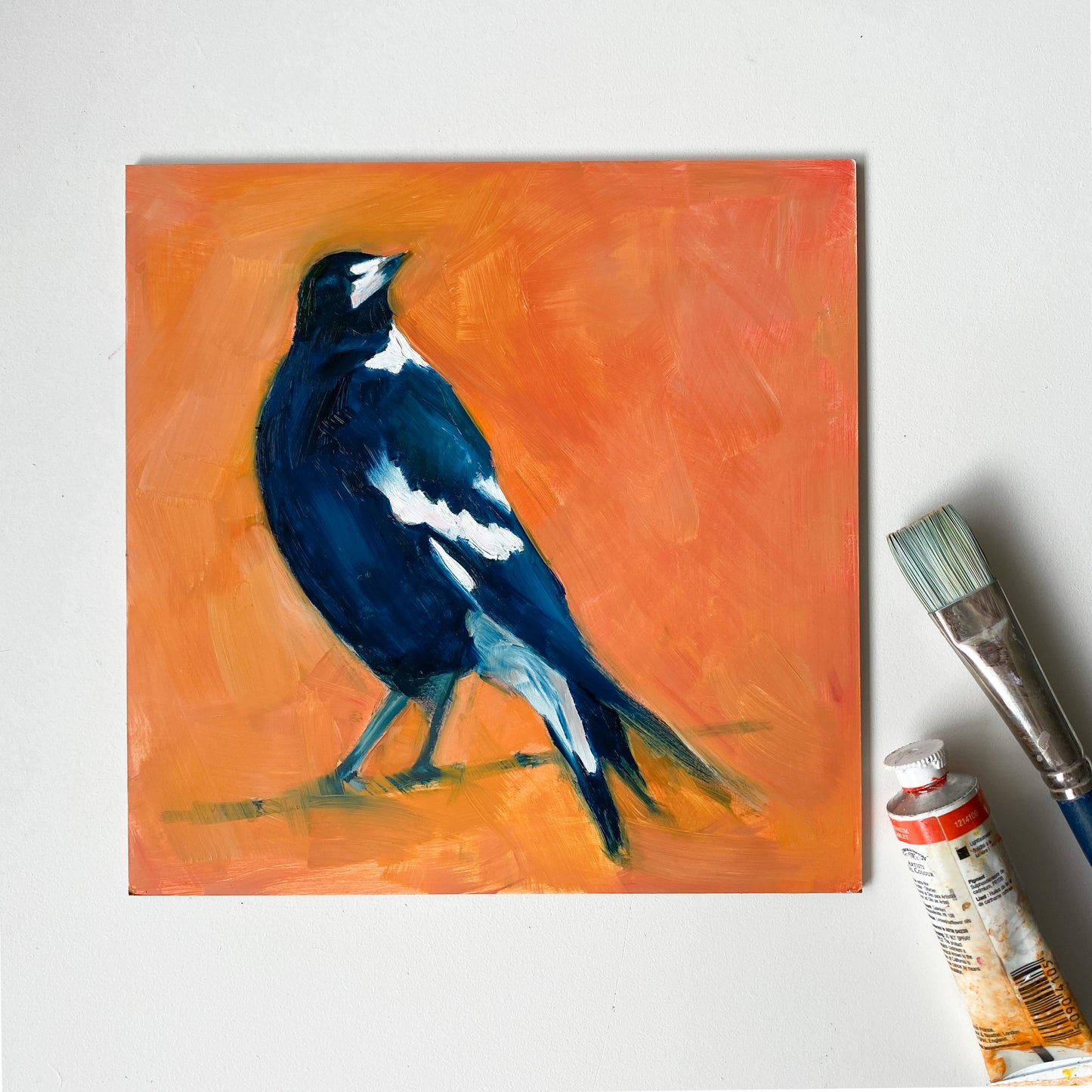 image of an impressionistic oil painting of a navy blue and white magpie looking up on a bright and textured orange background. the artwork is on a white table and there is a paint brush and oil paint tube next to it.