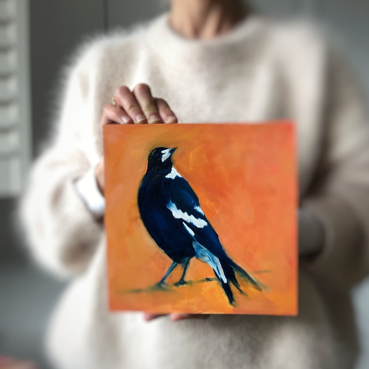 image of a person holding an impressionistic oil painting of a navy blue and white magpie looking up on a bright and textured orange background