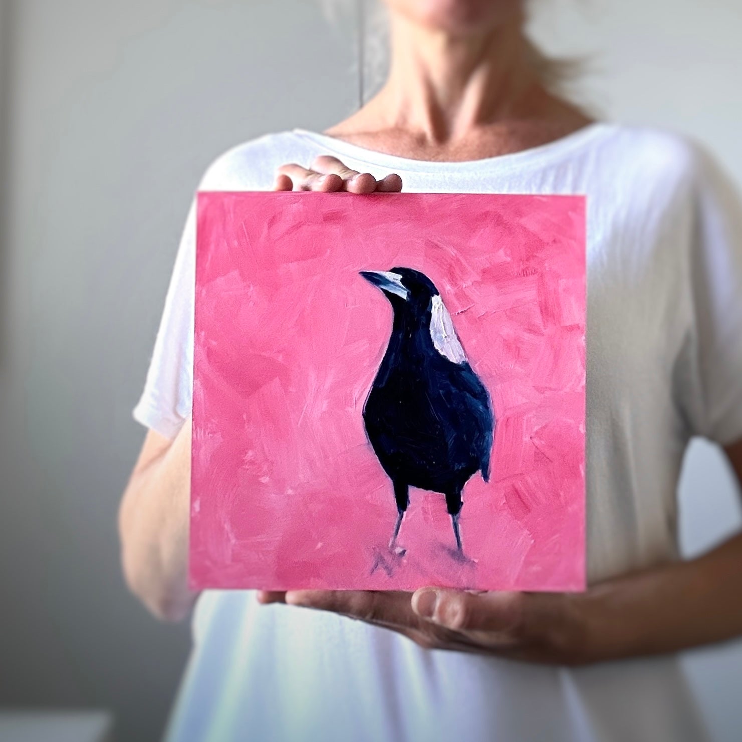 photo of a person holding a fine art original oil painting of a navy blue and white baby magpie on a textured pink background