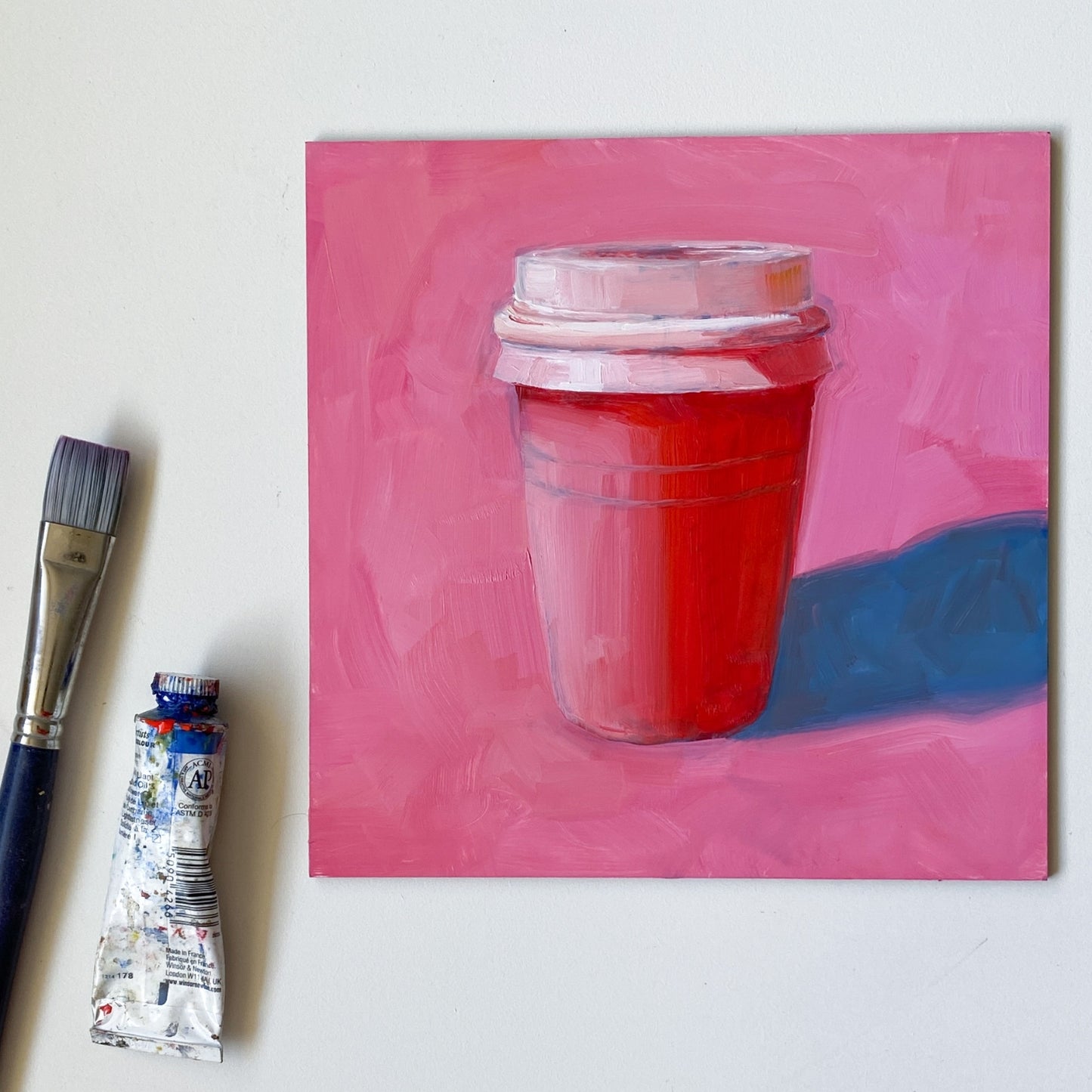 styled photo of an original oil painting of a pink and red take away coffee cup on a textured bright pink background with strong greyish blue shadows. The painting is on a white desk and there's a paintbrush and oil paint tube next to it.