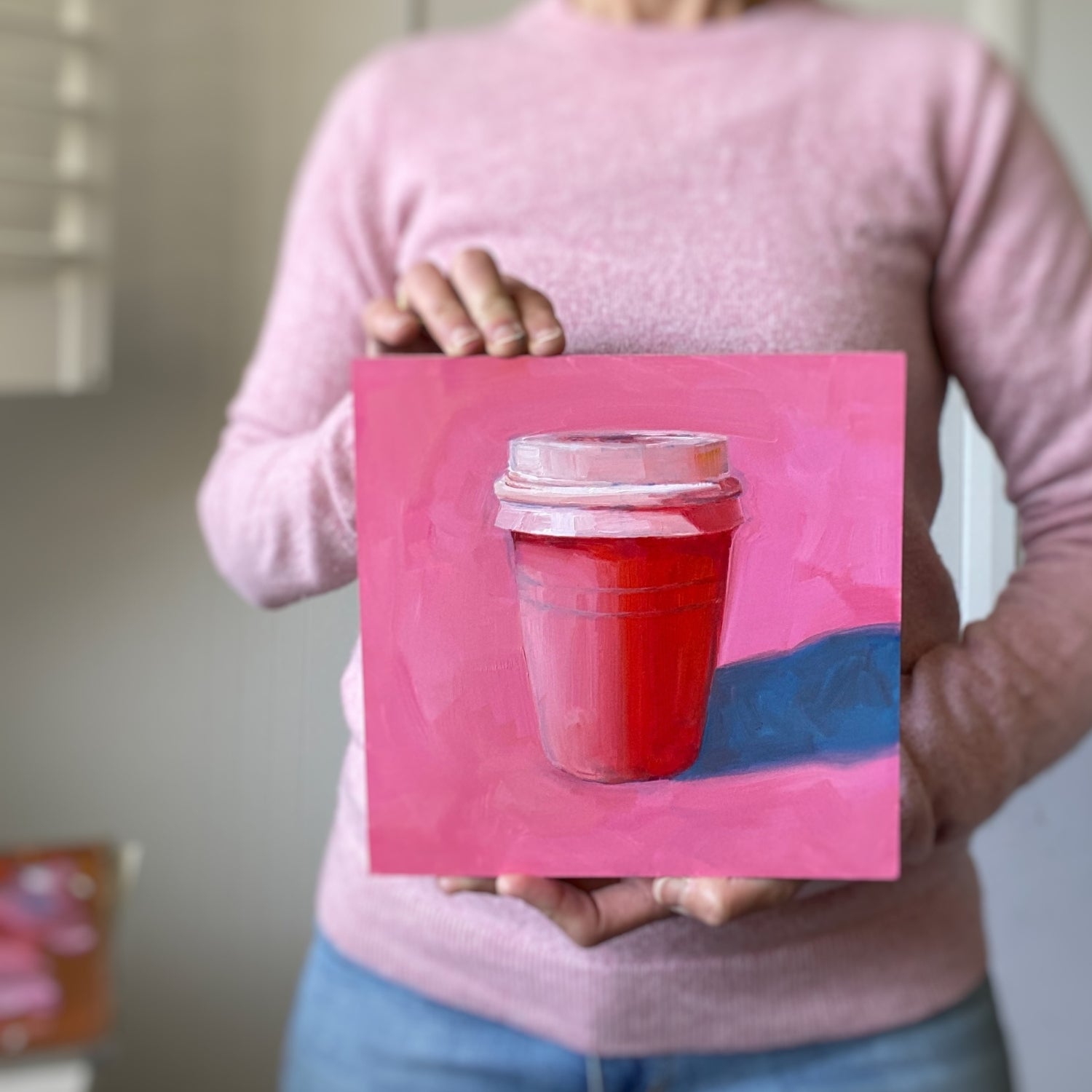 photo of a person holding an original oil painting of a pink and red take away coffee cup on a textured bright pink background with strong greyish blue shadows.