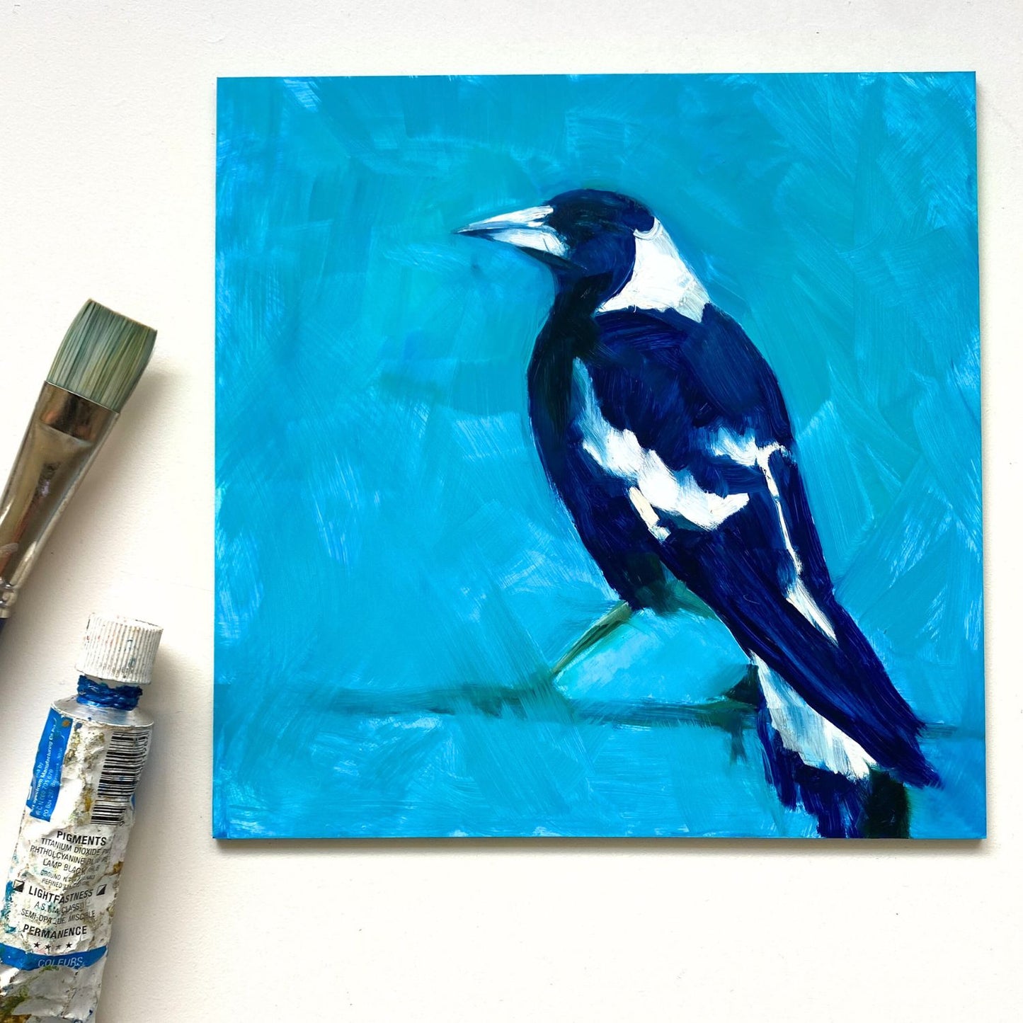 styled photo of an original oil painting of the profile of a navy blue and white magpie on a textured turquoise background. The painting is on a white desk and there's a paintbrush and oil paint tube next to it.