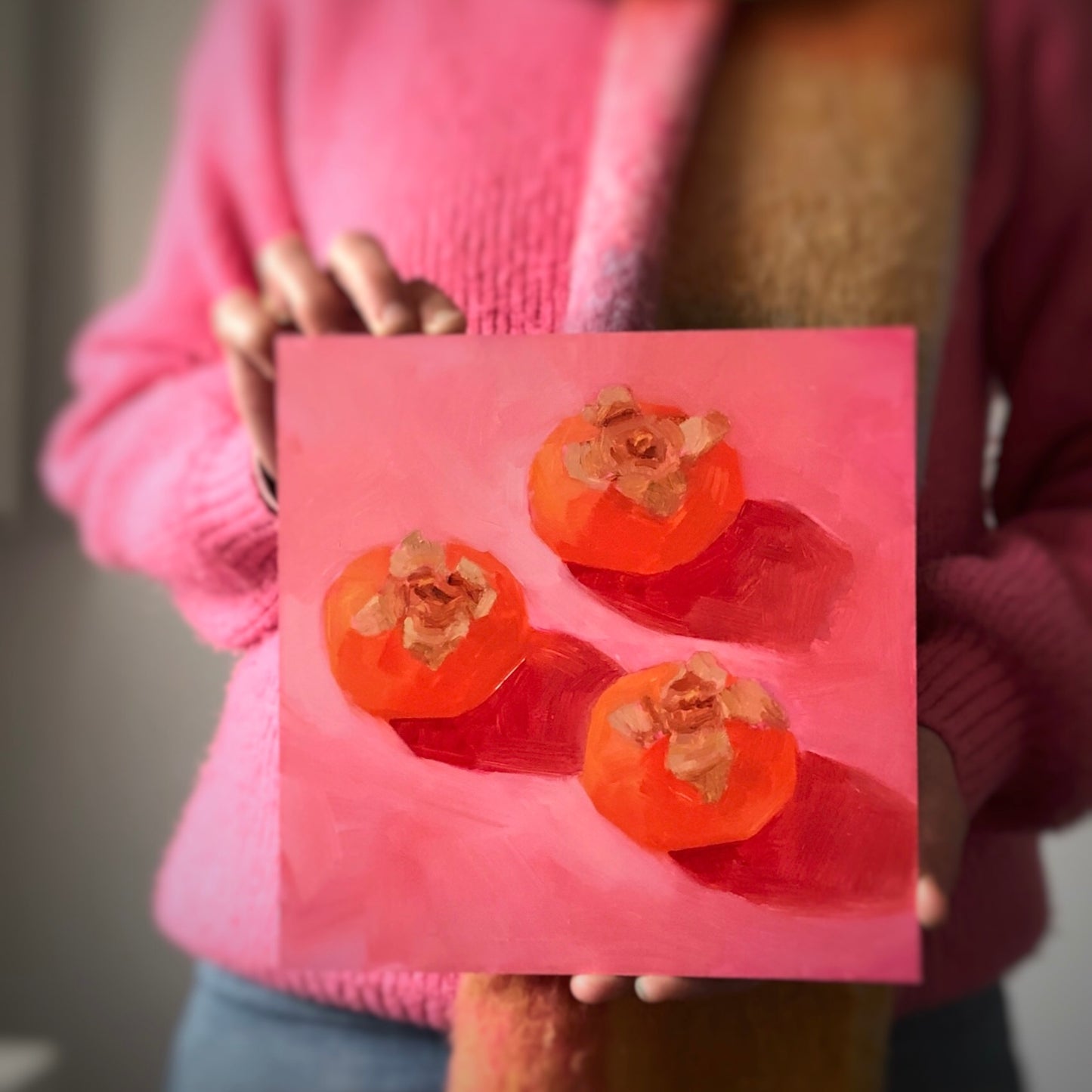 image of a person holding an original oil painting of three orange persimmons on soft pink with strong shadows