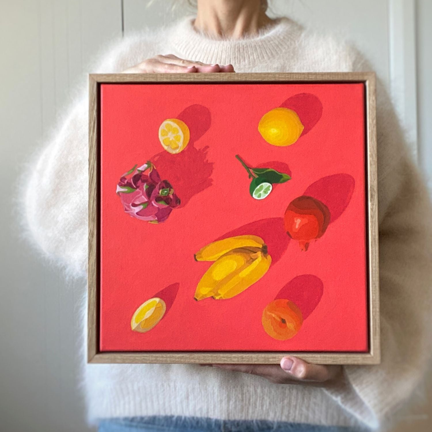 photo of a person holding an original oil painting of fresh fruits like bananas, lemons, dragonfruit, pomegranate, lime and apricot on a vibrant and bright red background with beautiful shadows