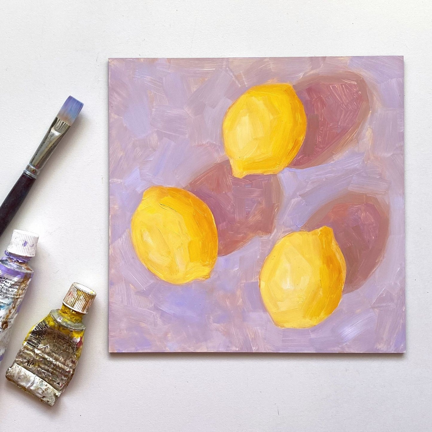 lifestyle photo of an original oil painting of three vibrant lemons with strong dark pink shadows on a textured lilac background. the artwork is on a white desk and there is a paintbrush and oil tubes next to it