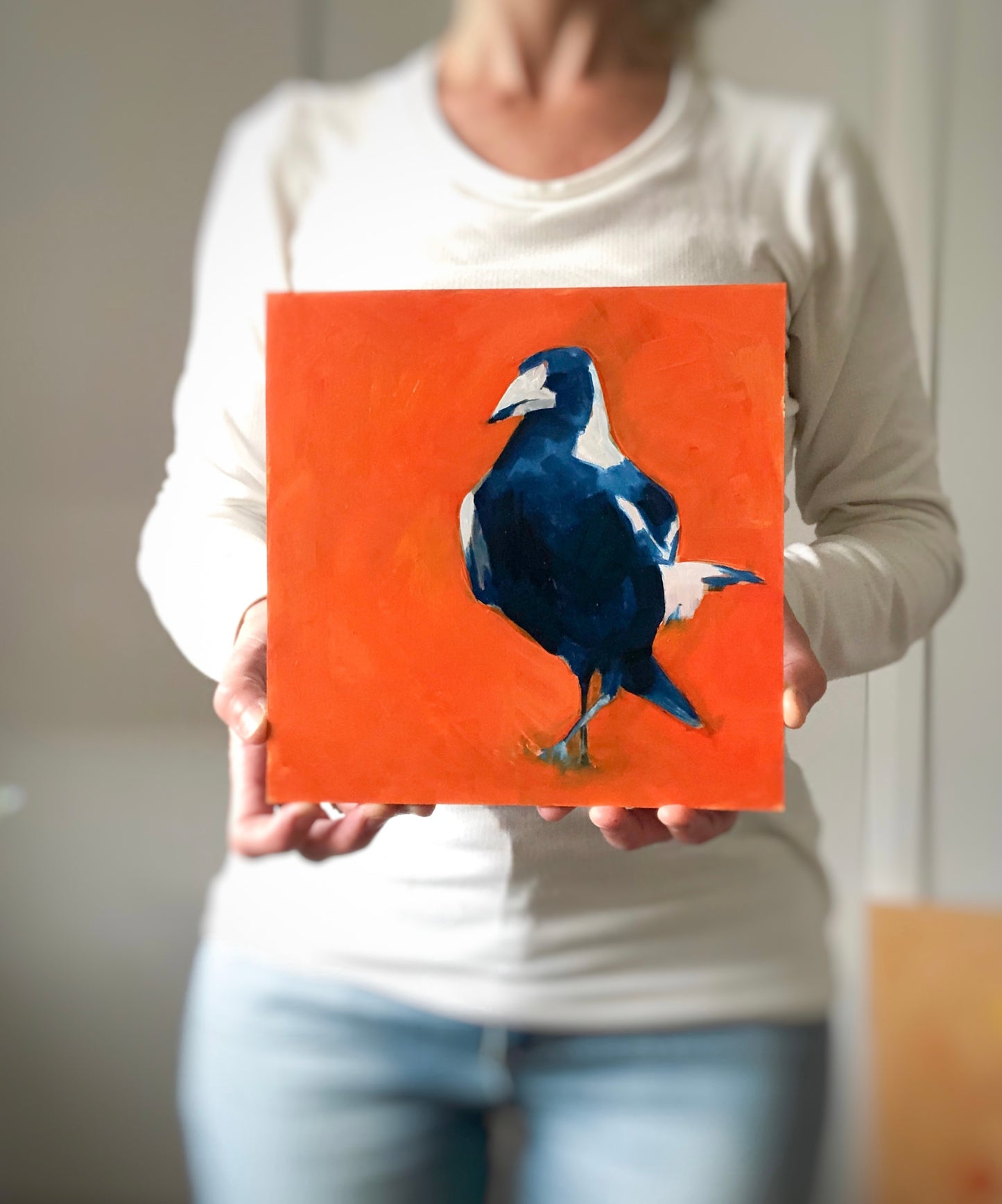 photo of a woman holding an oil painting on panel closeup of the head of a magpie in blue with a bright orange background