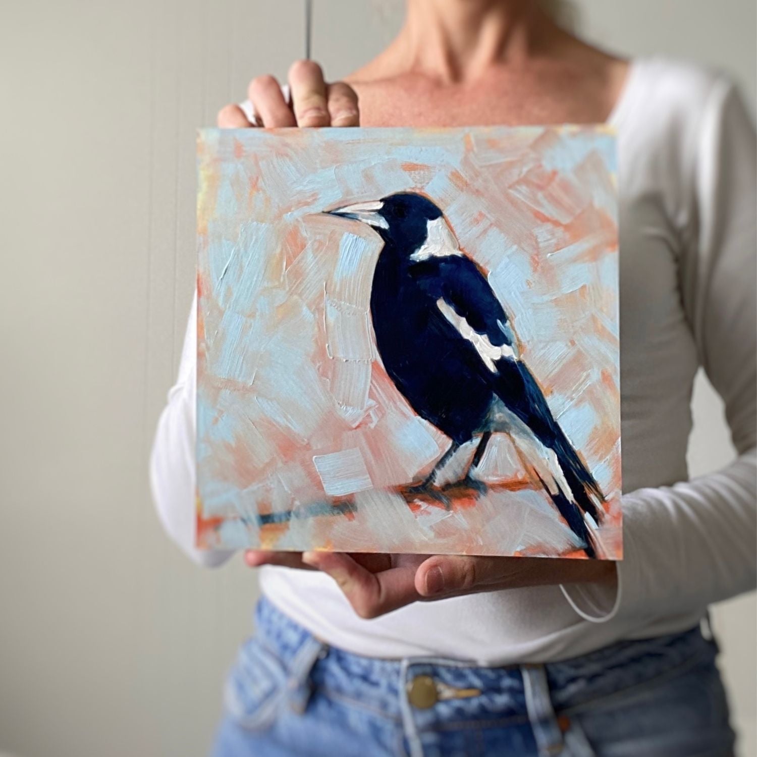 photo of a person holding an original oil painting of a navy blue and white magpie on a textured sky blue background where you can see orange coming through
