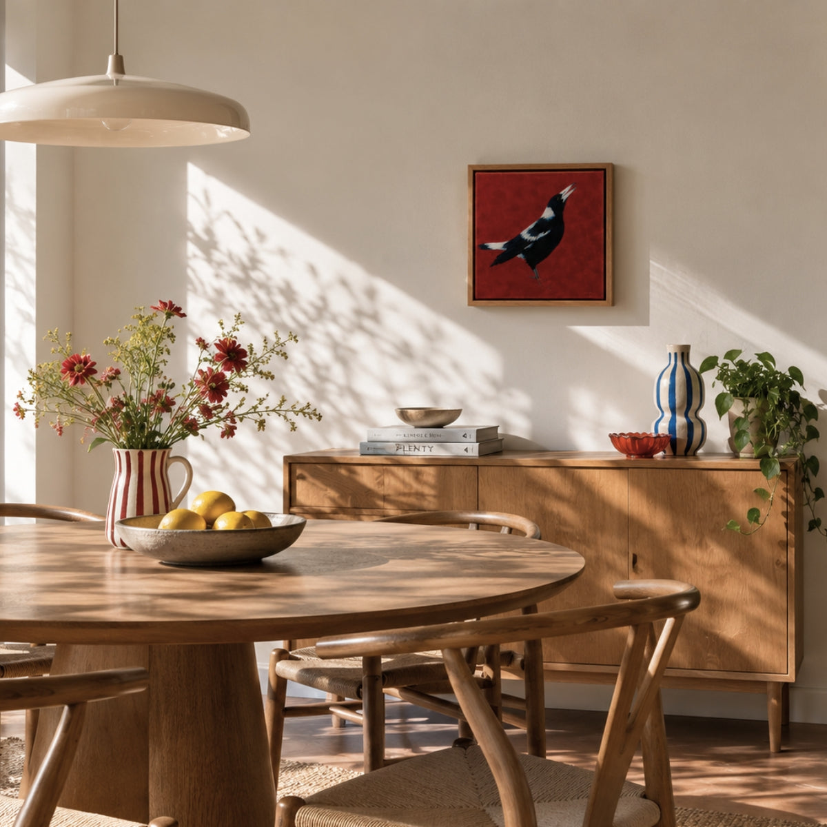 A framed art print featuring a magpie on a textured red background, styled on a light beige wall in a warm, sunlit dining room with a wooden table, chairs, and sideboard.