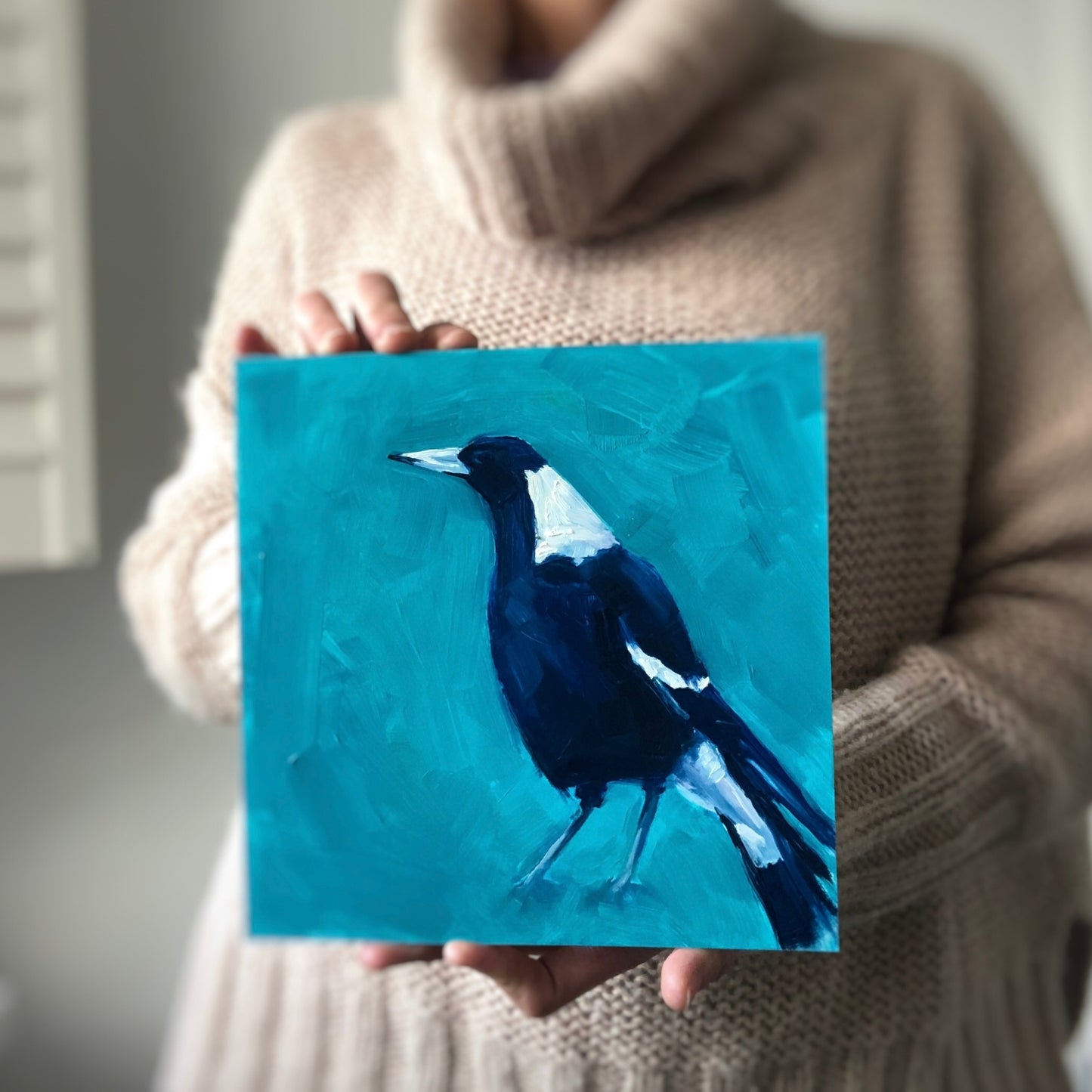 photo of person holding an original oil painting on board of a navy blue and white magpie on a textured turquoise background