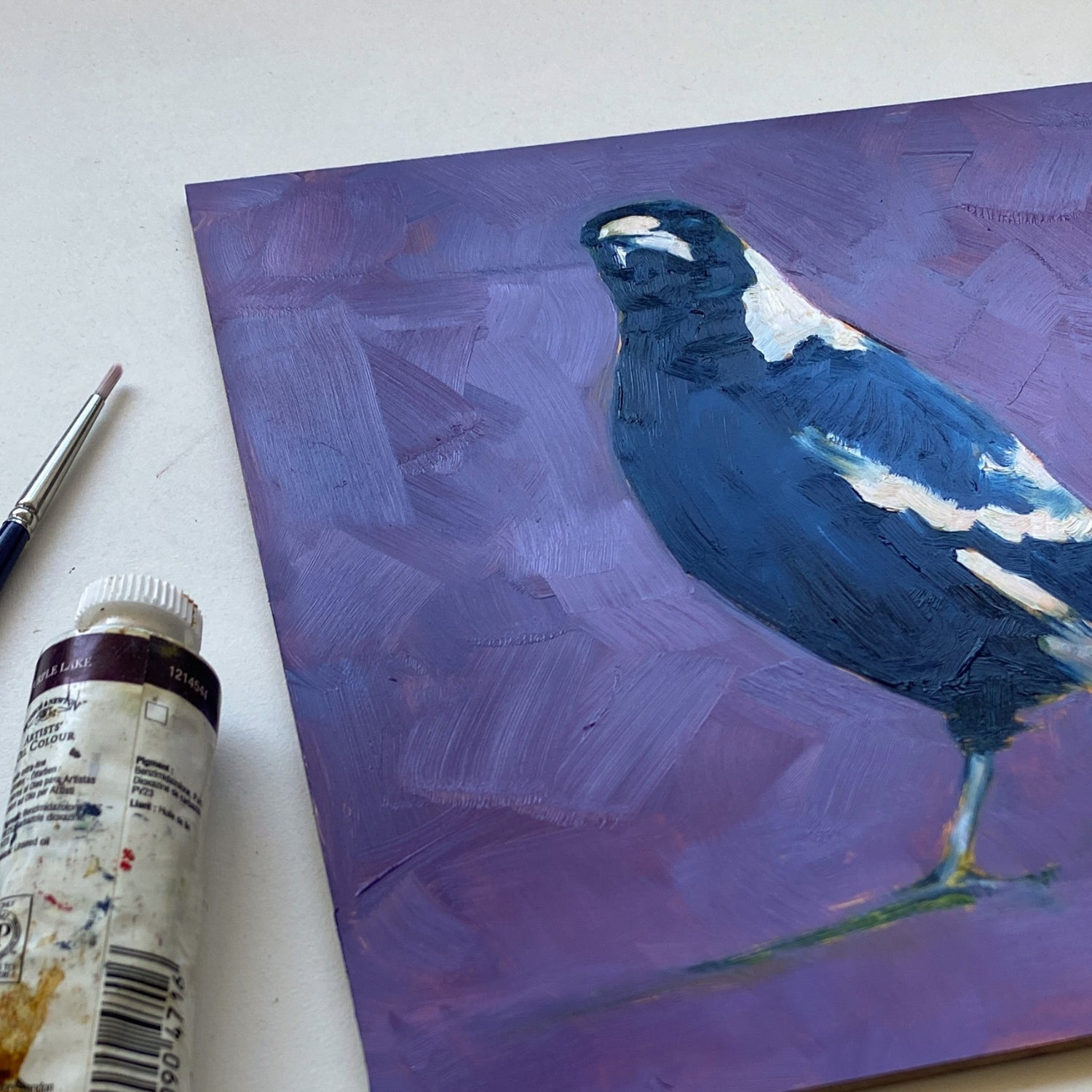 styling photo of a closeup of an original oil painting of a navy blue and white magpie with its head tilted on a bright and textured purple with bits of blue background. The painting is sitting on a white surface and there is a paintbrush and paint tube next to it.