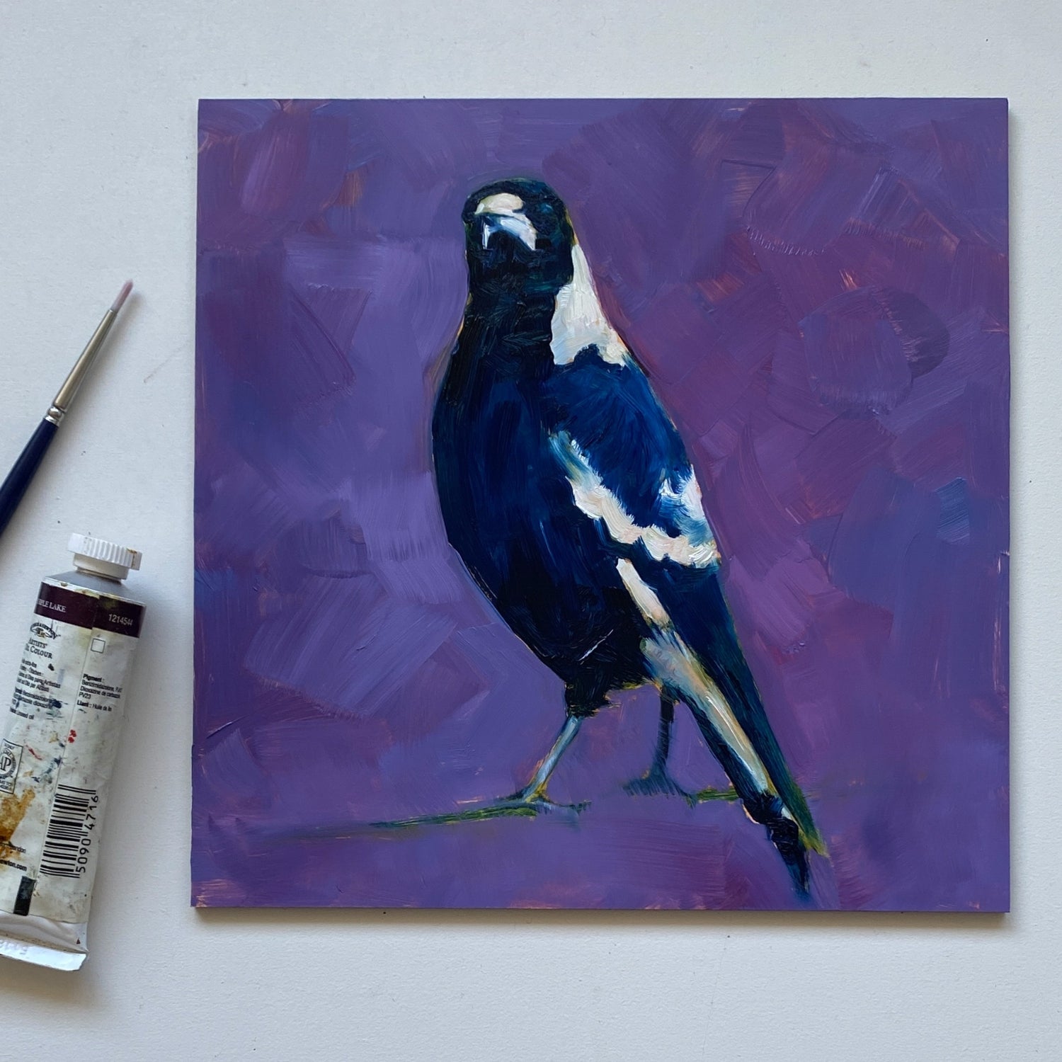 styling photo of a closeup of an original oil painting of a navy blue and white magpie with its head tilted on a bright and textured purple with bits of blue background. The painting is sitting on a white surface and there is a paintbrush and paint tube next to it.