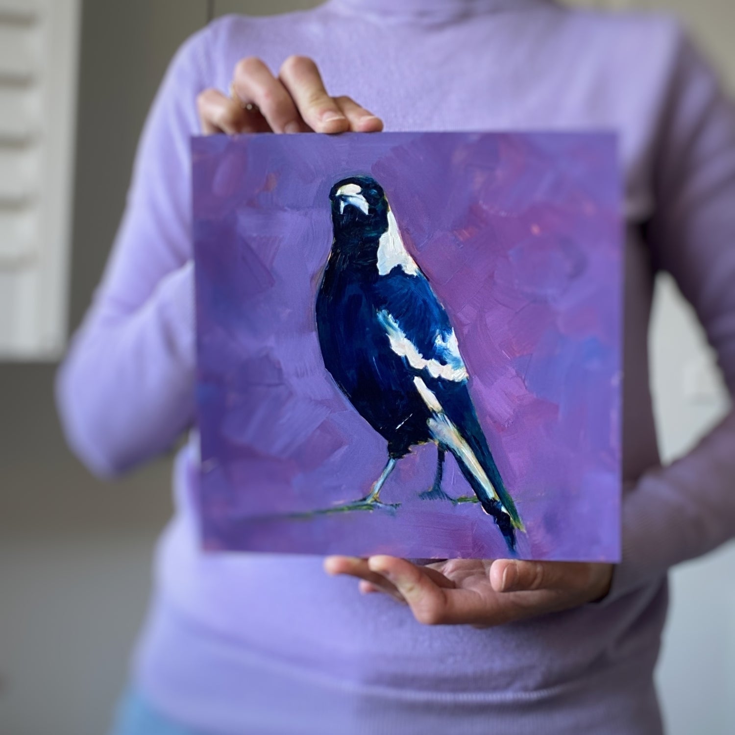 lifestyle photo of a person holding an original painting of a navy blue and white magpie with its head tilted on a bright and textured purple with bits of blue background.