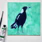 lifestyle photo of an original oil painting of a navy blue and white magpie on a textured minty green background. the painting is on a white desk and there is a paintbrush and oil paint tube next to it