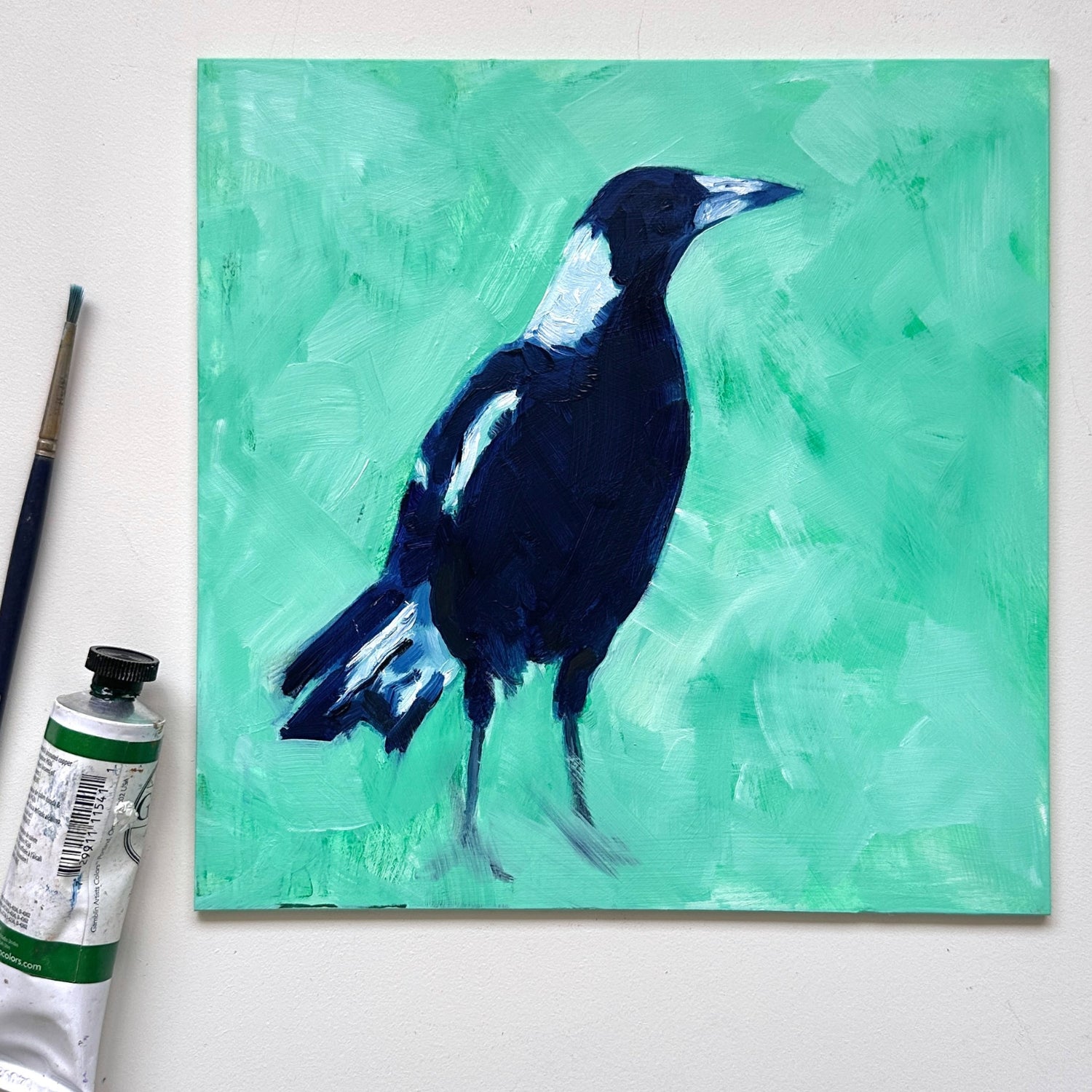lifestyle photo of an original oil painting of a navy blue and white magpie on a textured minty green background. the painting is on a white desk and there is a paintbrush and oil paint tube next to it