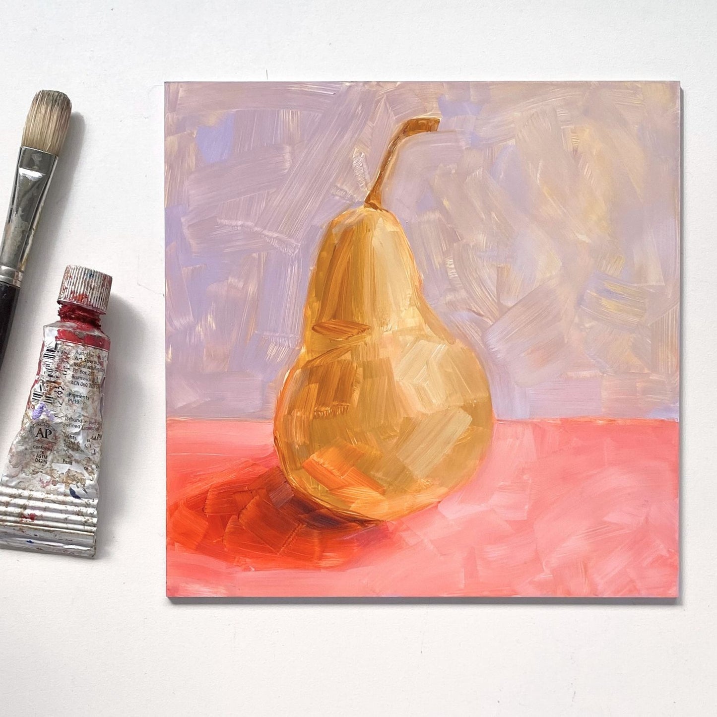 lifestyle image of an original oil painting of a yellow / beige pear on a textured pink surface and lilac background. the painting is on a white desk and there is a paintbrush and oil paint tube next to it