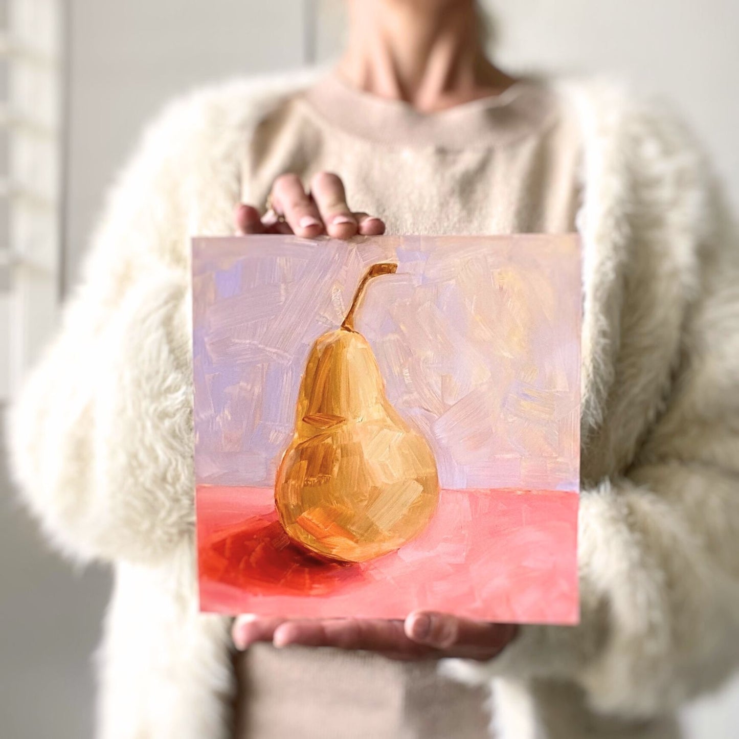 photo of a person holding an original oil painting of a yellow / beige pear on a textured pink surface and lilac background