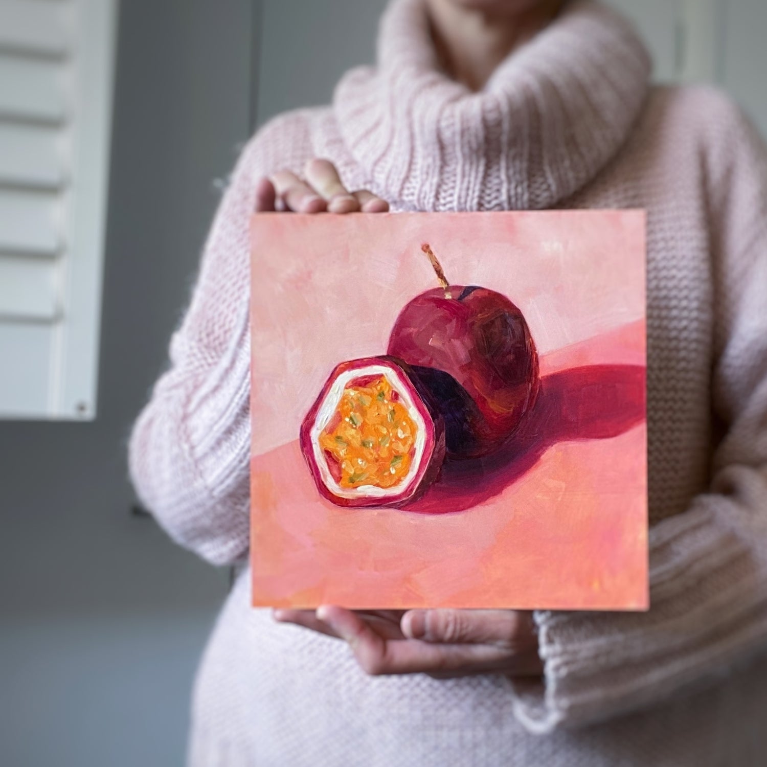 lifestyle photo of a person holding an original painting of a burgundy, magenta whole passionfruit and half a passionfruit with seeds. They are sitting on a soft pink surface and there are strong shadows and a diagonal background. 