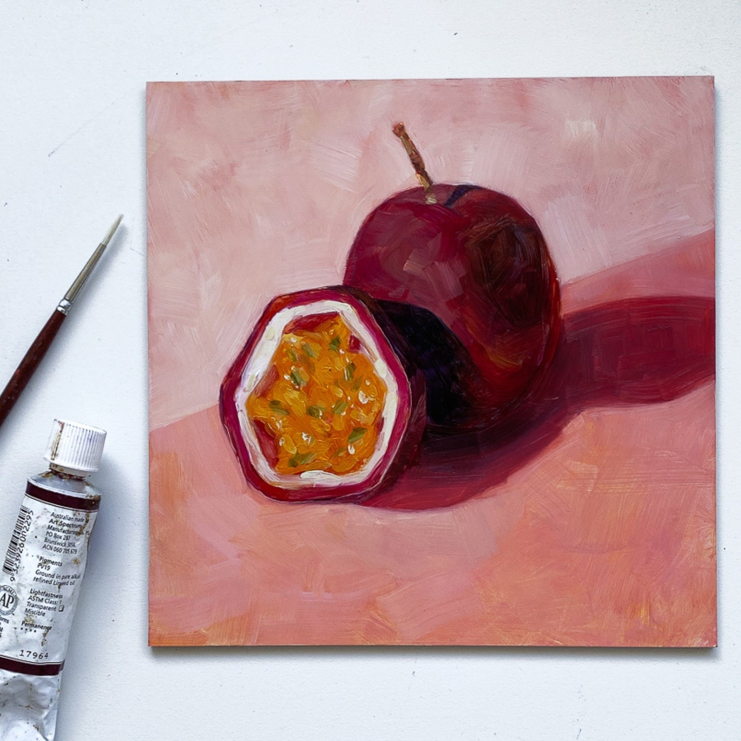 styling photo of an original painting of a burgundy, magenta whole passionfruit and half a passionfruit with seeds. They are sitting on a soft pink surface and there are strong shadows and a diagonal background. The painting is on a white surface and there is a paintbrush and oil paint tube next to it.
