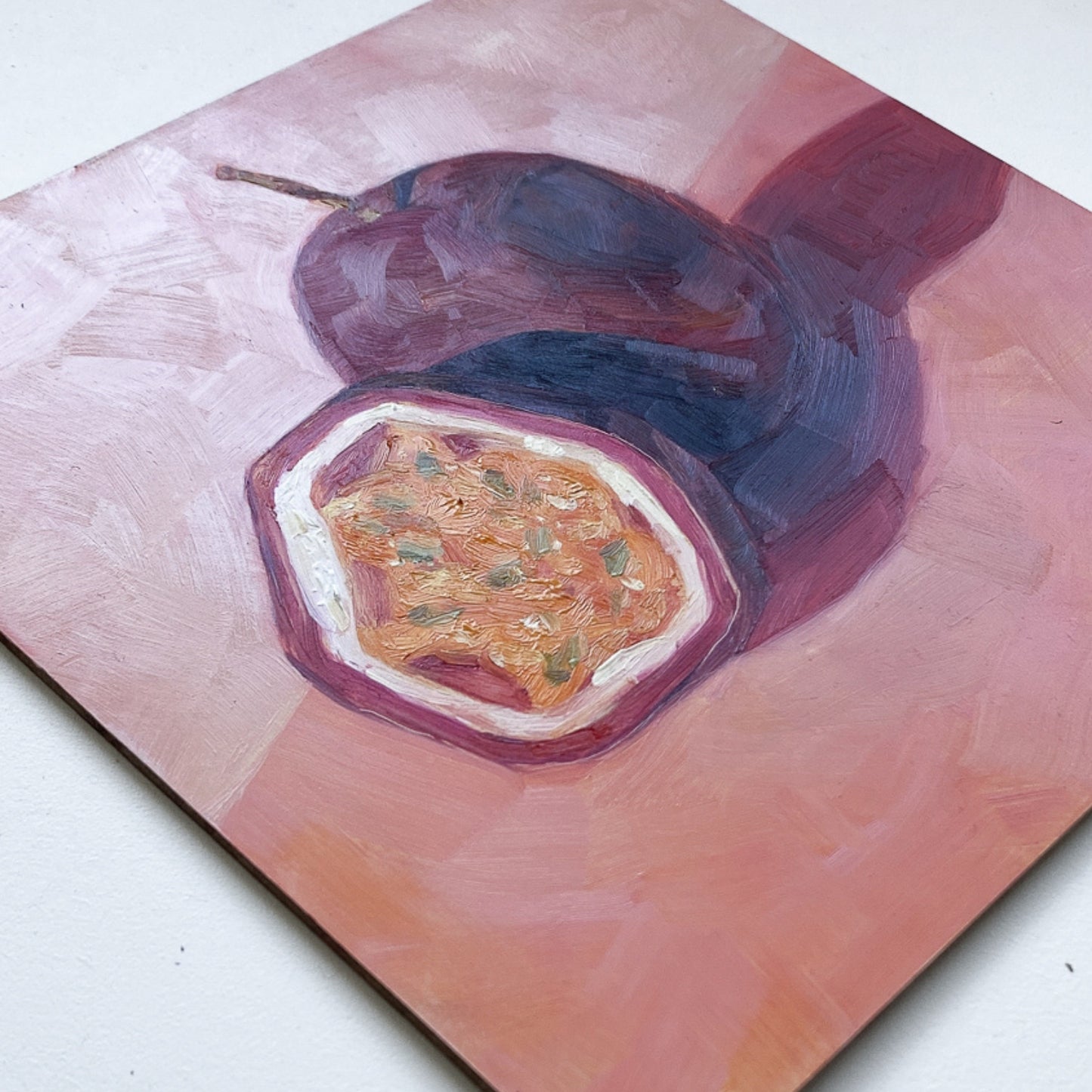 an original painting of a burgundy, magenta whole passionfruit and half a passionfruit with seeds. They are sitting on a soft pink surface and there are strong shadows and a diagonal background. The painting is on a white surface.
