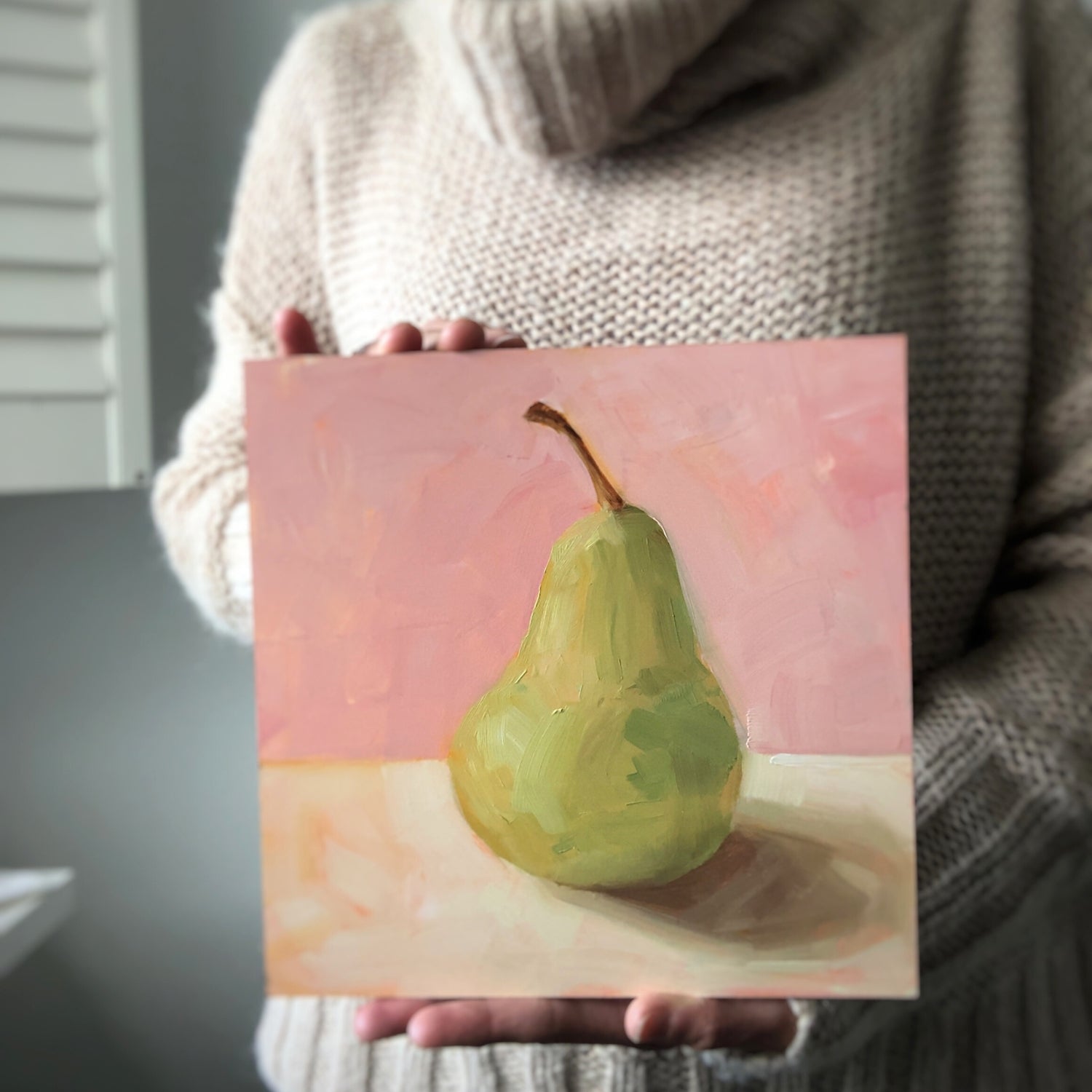 photo of a person holding an impressionistic and realistic original oil painting on board of a green pear sitting on a soft pink and cream background