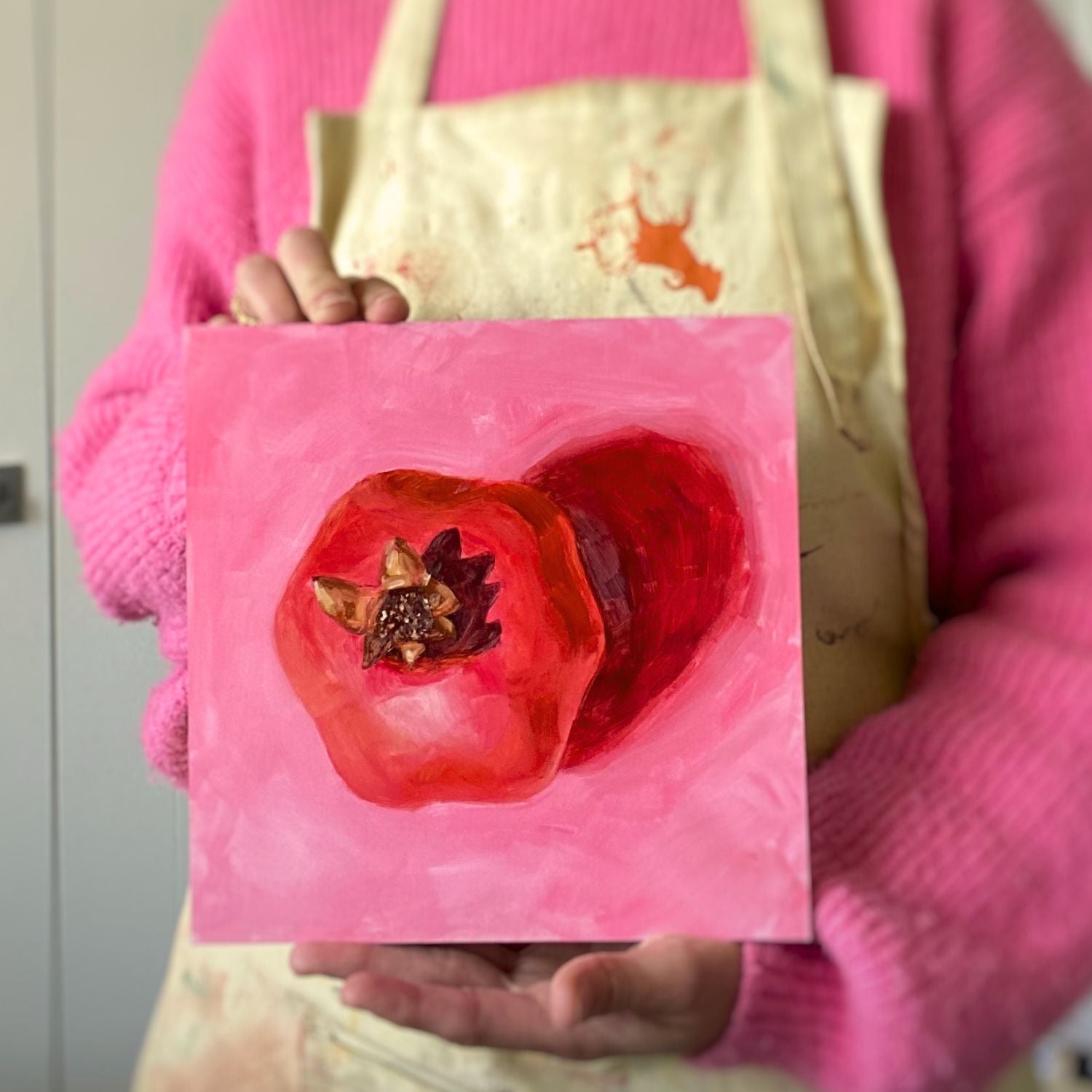photo of a person holding an original oil painting of a bright and vibrant red pomegranate with a strong reddish shadow on a textured pink background