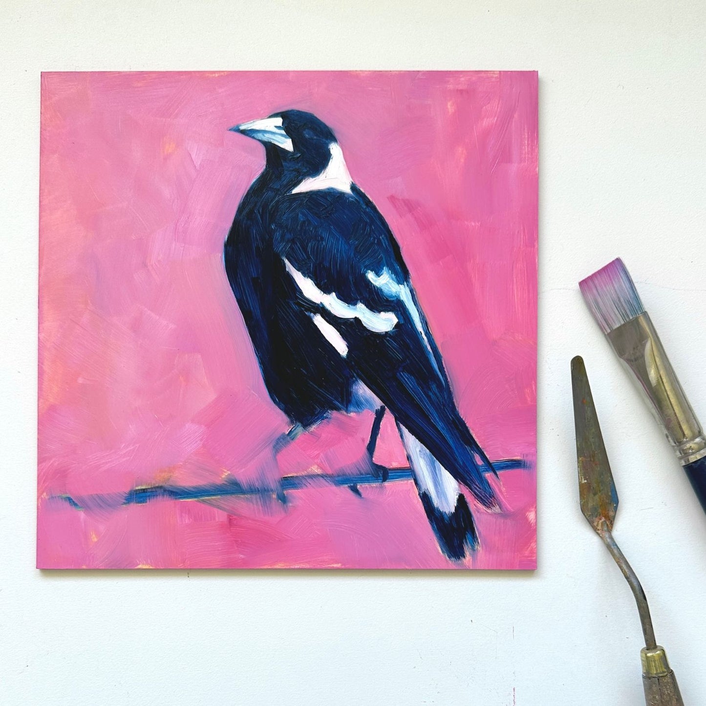 lifestyle photo of an original oil painting of a navy blue and white magpie on a textured bright pink background. the painting is on a white desk and there is a paintbrush and palette knife next to it