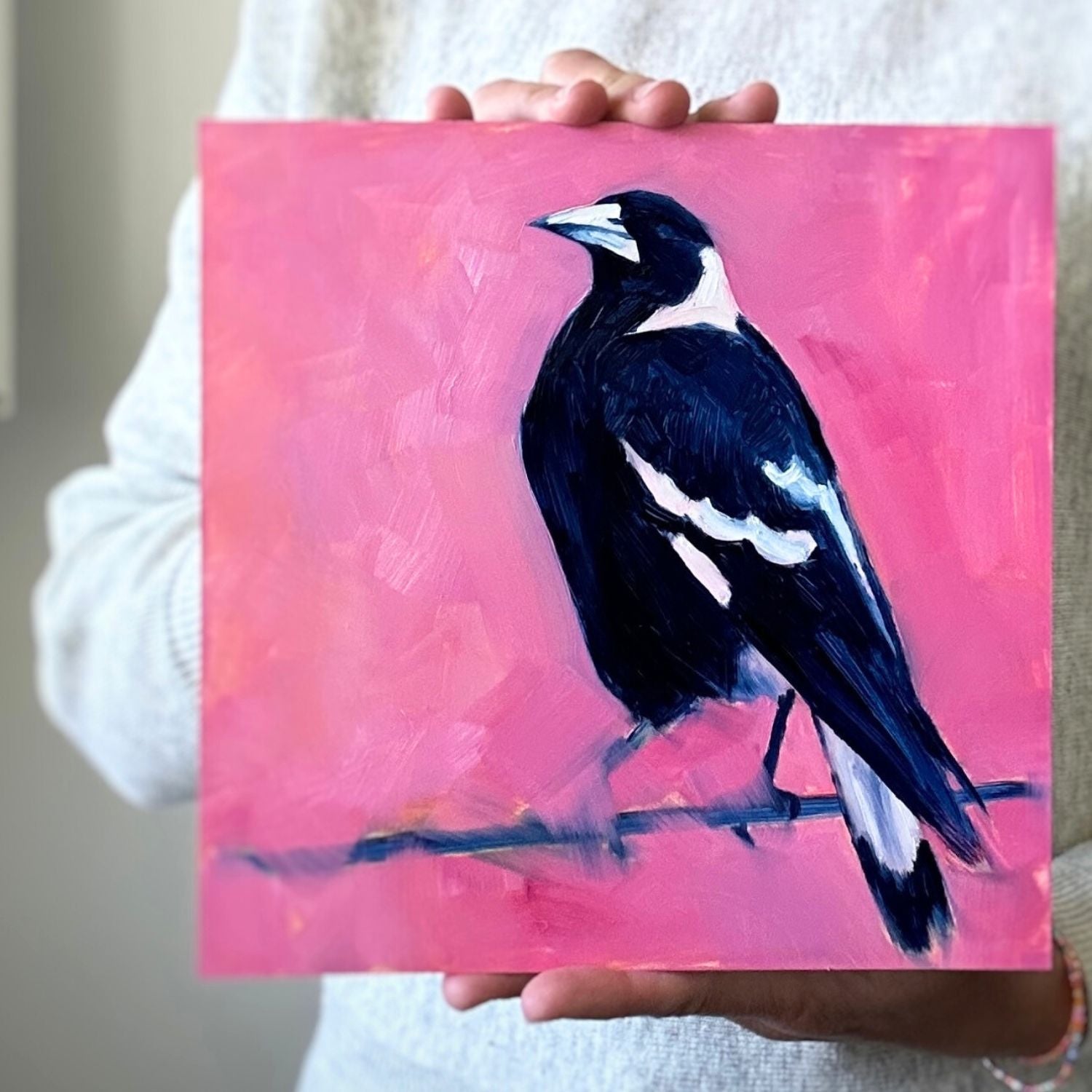 photo of a person holding an original oil painting of a navy blue and white magpie on a textured bright pink background 