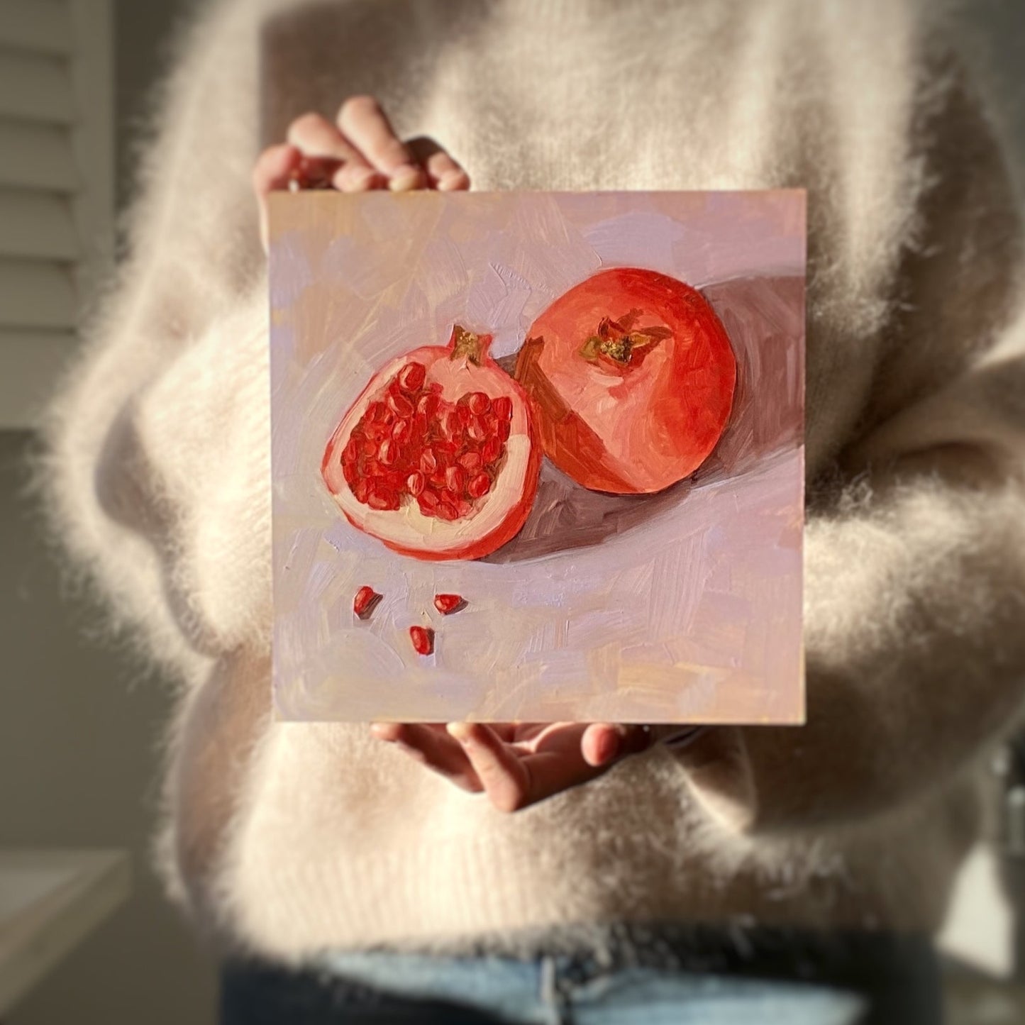 image of a person holding an original oil painting of a whole pomegranate and a cut pomegranate with seeds lying on a soft and smooth lilac background with bits of yellow showing through