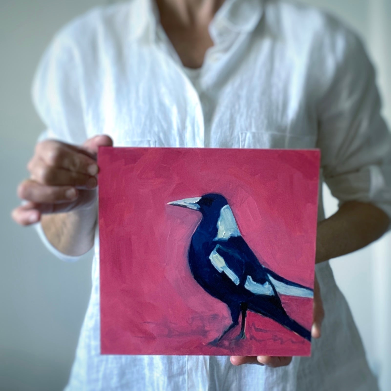 photo of a person holding an oil painting on panel of a magpie in navy blue and white on a bright pink background
