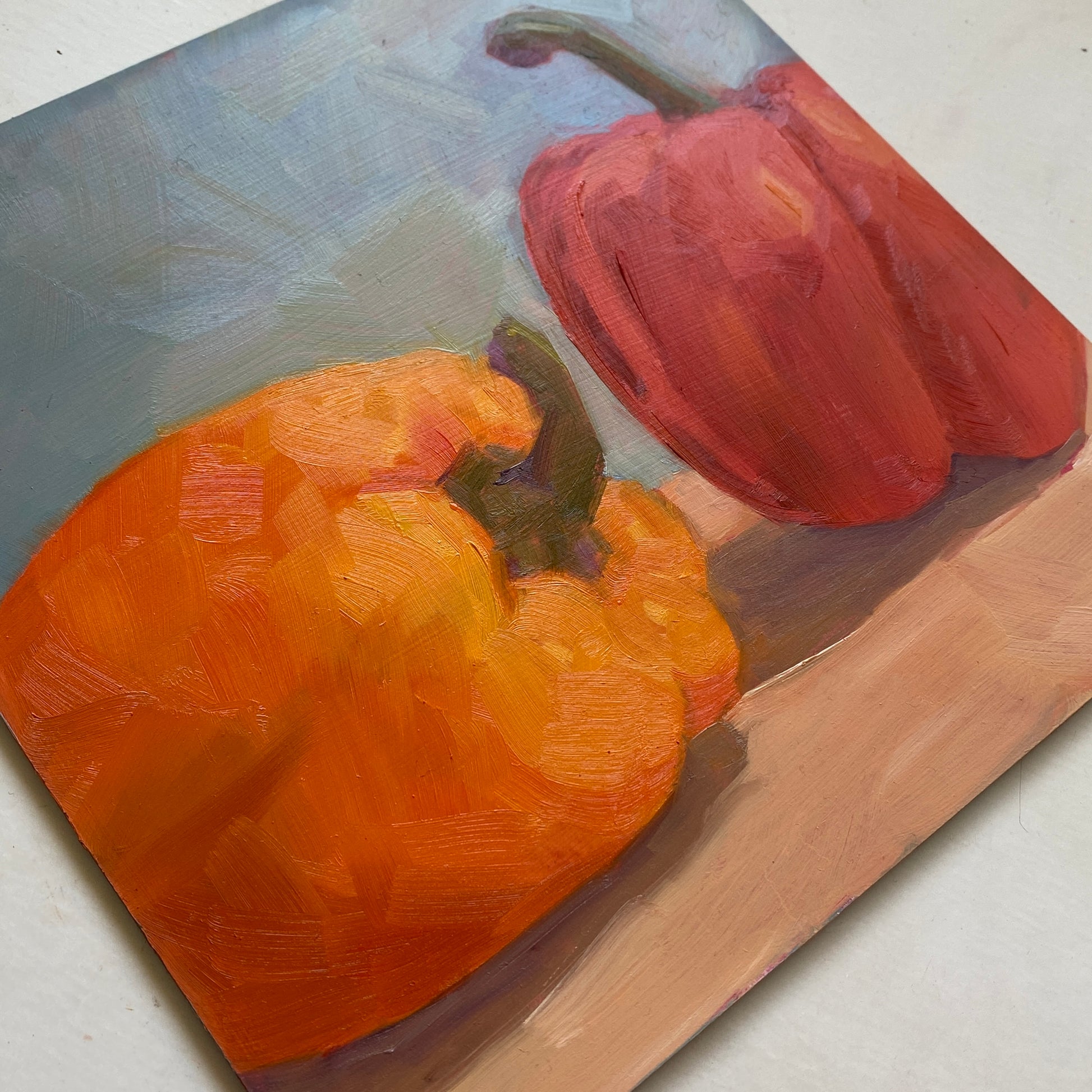 an image of red and orange capsicum oil painting on a wooden panel with light grey-blue background and beige table