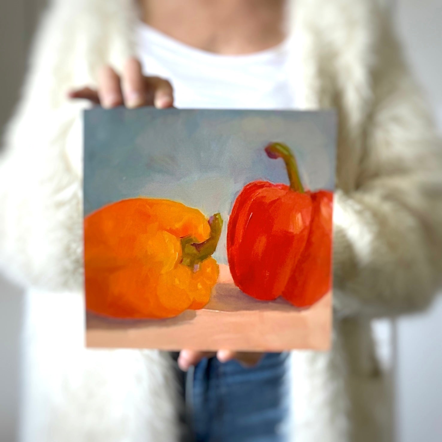 a photo of a person holding a red and orange capsicum oil painting on a wooden panel with light grey-blue background and beige table