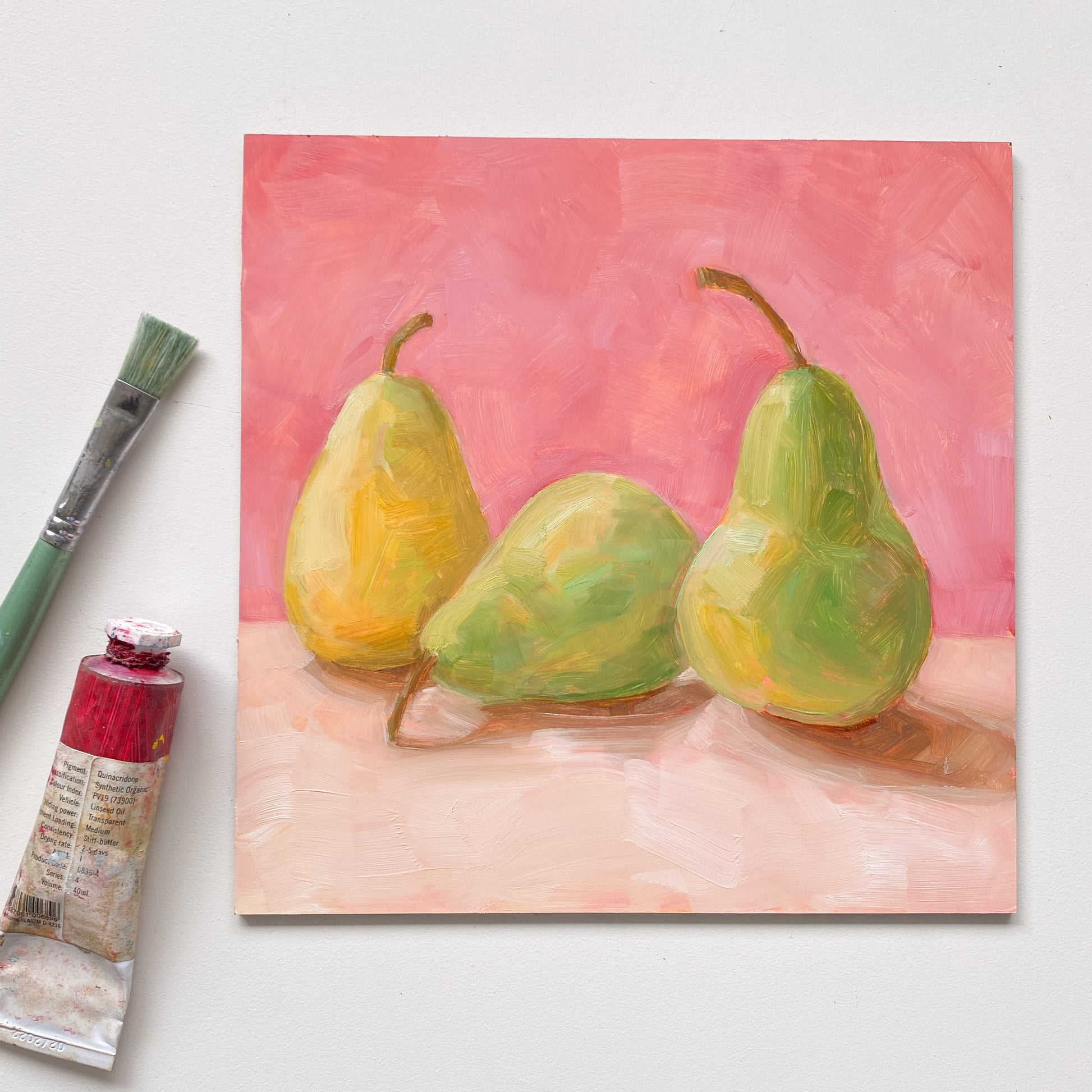 image of an oil painting of two green pears and one yellow pear on a cream surface with a soft and warm pink background. the artwork is on a white table and there is a paint brush and oil paint tube next to it