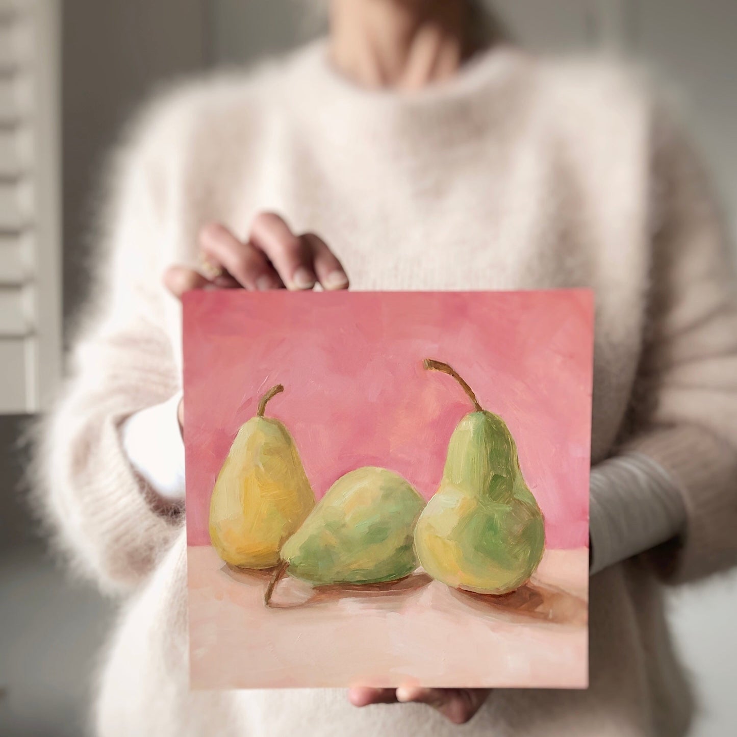 image of a person holding an oil painting of two green pears and one yellow pear on a cream surface with a soft and warm pink background
