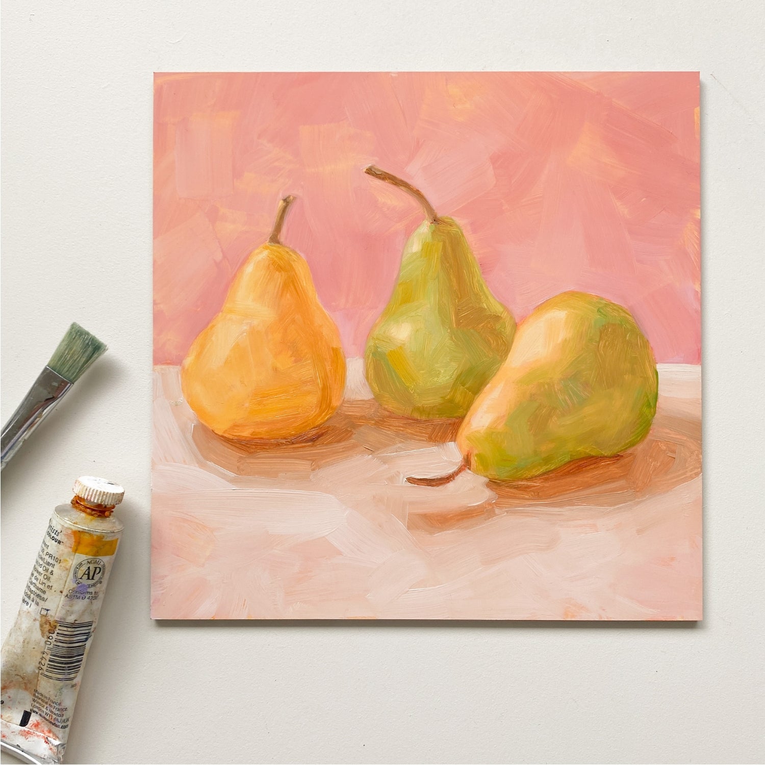 styled photo of an original oil painting of three yellow and green pears on a soft cream surface and a soft pink background painted in an impressionistic way. The painting is on a white desk and there is a paintbrush and a paint tube next to it
