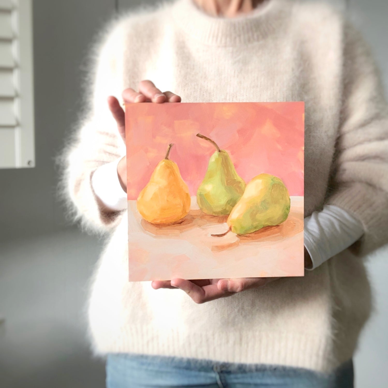photo of a person holding an original oil painting of three yellow and green pears on a soft cream surface and a soft pink background painted in an impressionistic way