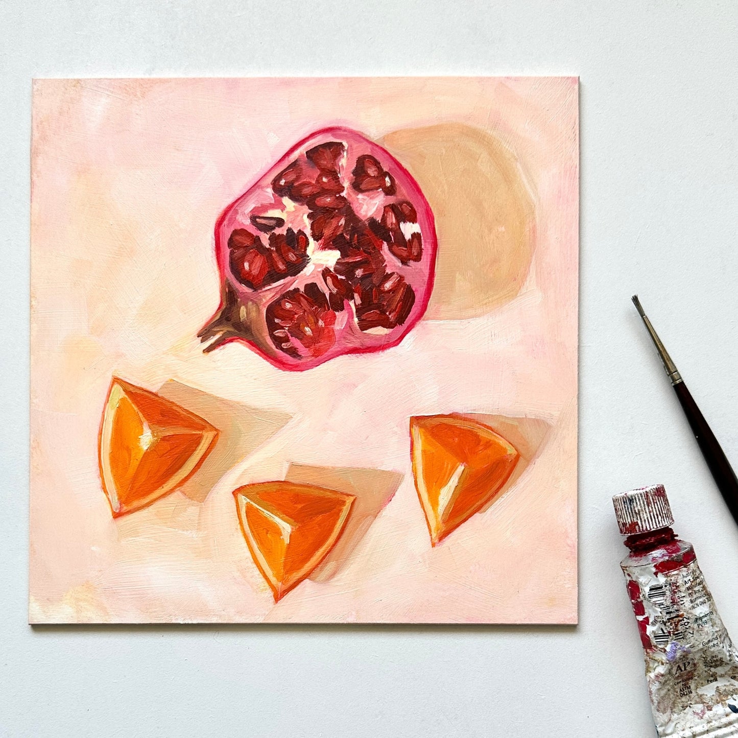 lifestyle photo impressionistic original oil painting of a still life setting of pomegranate fruit and three orange slices on a textured cream background with soft shadows. the painting is on a white desk and there is a paintbrush and oil paint tube next to it