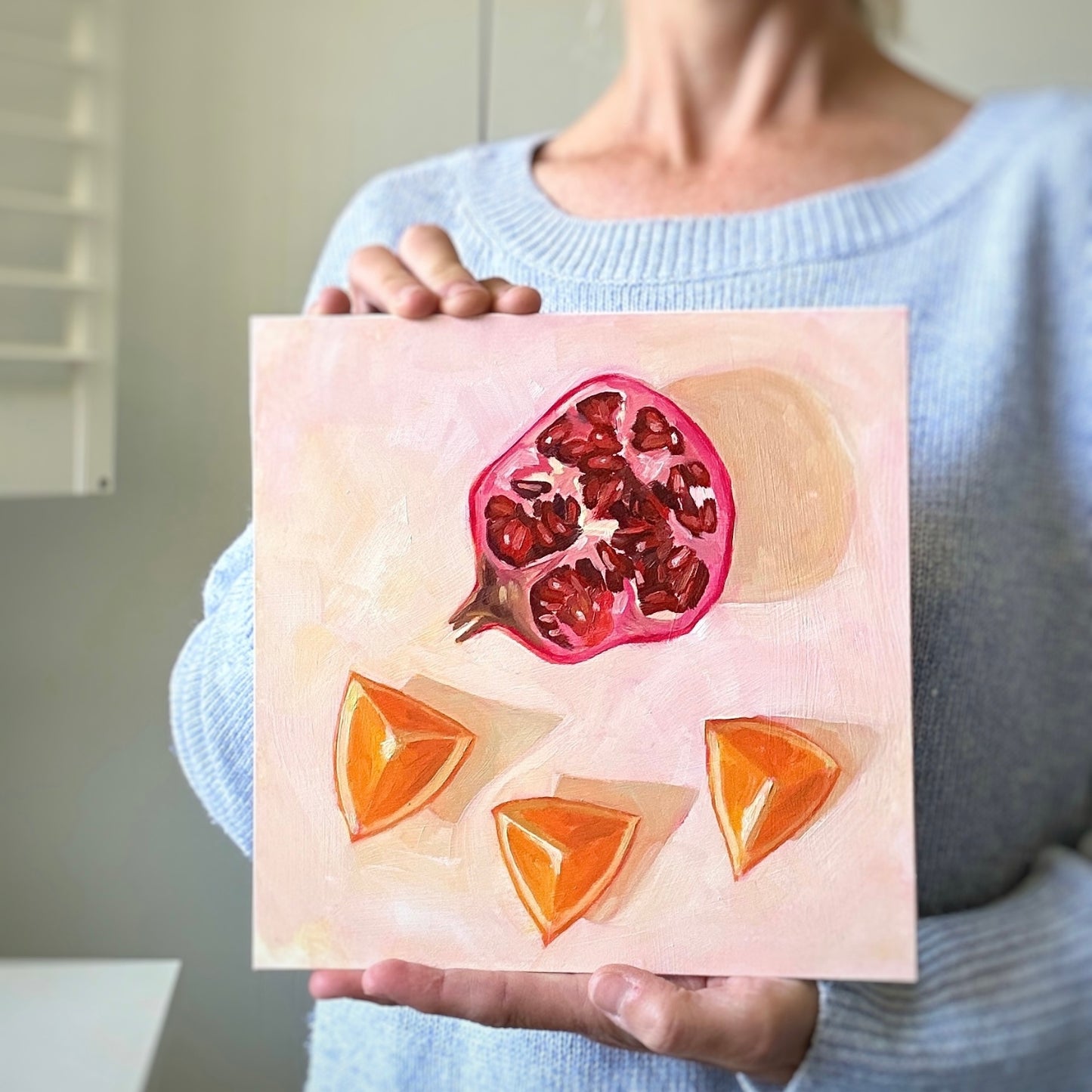 photo of a person holding an impressionistic original oil painting of a still life setting of pomegranate fruit and three orange slices on a textured cream background with soft shadows
